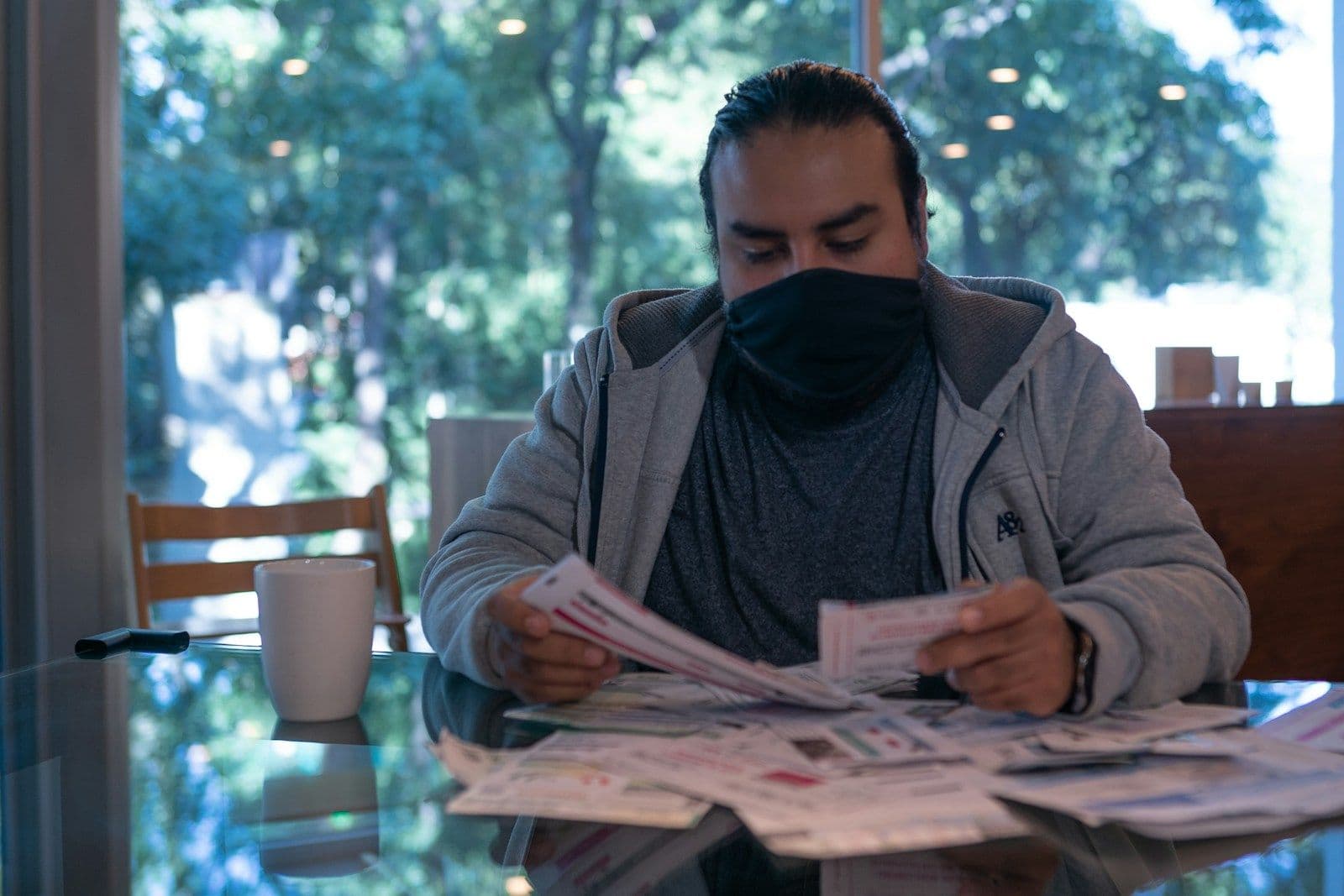 A man wearing a black face mask and gray hoodie sits at a glass table sorting through a stack of paper statements and bills, with a white mug nearby and trees visible through the window.