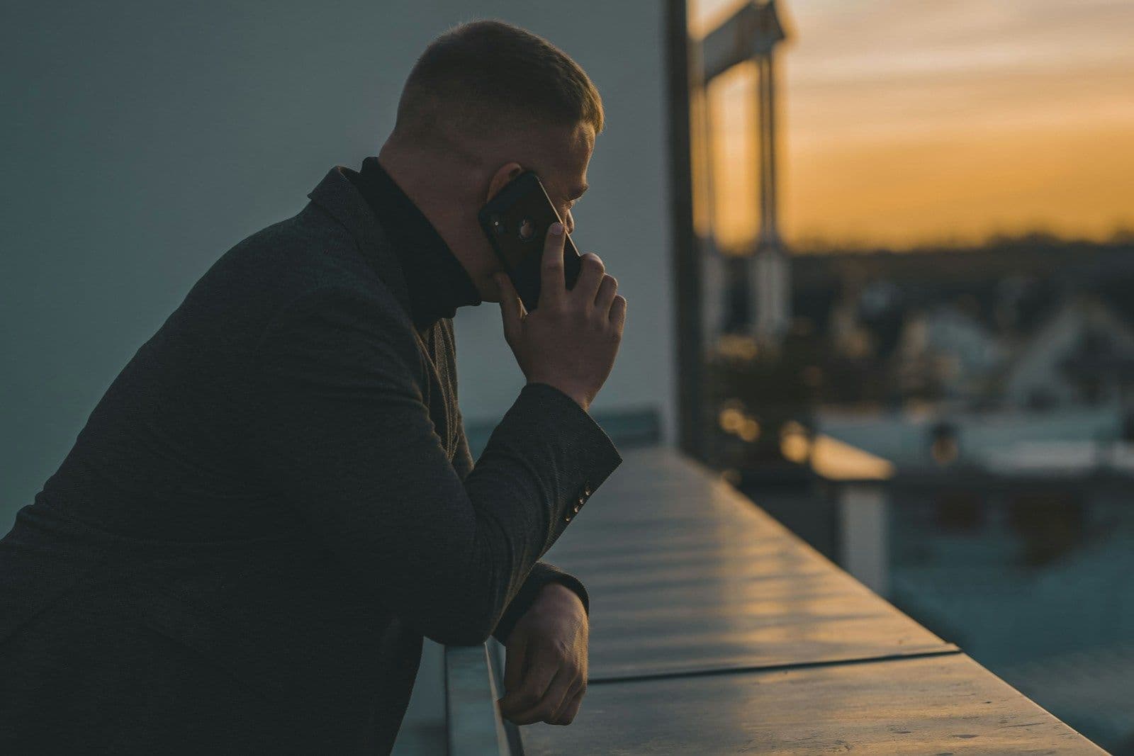 Man in a dark jacket leaning on a balcony railing at sunset, holding a phone to his ear and looking out over a city view
