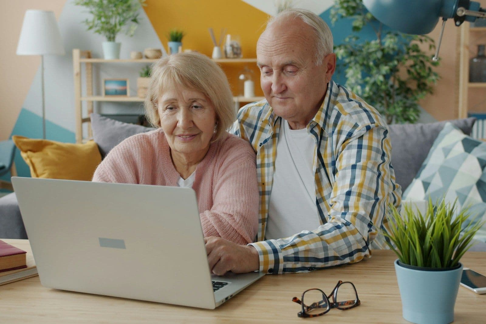 An older couple sits at a table in a bright living room, looking closely at a laptop together.
