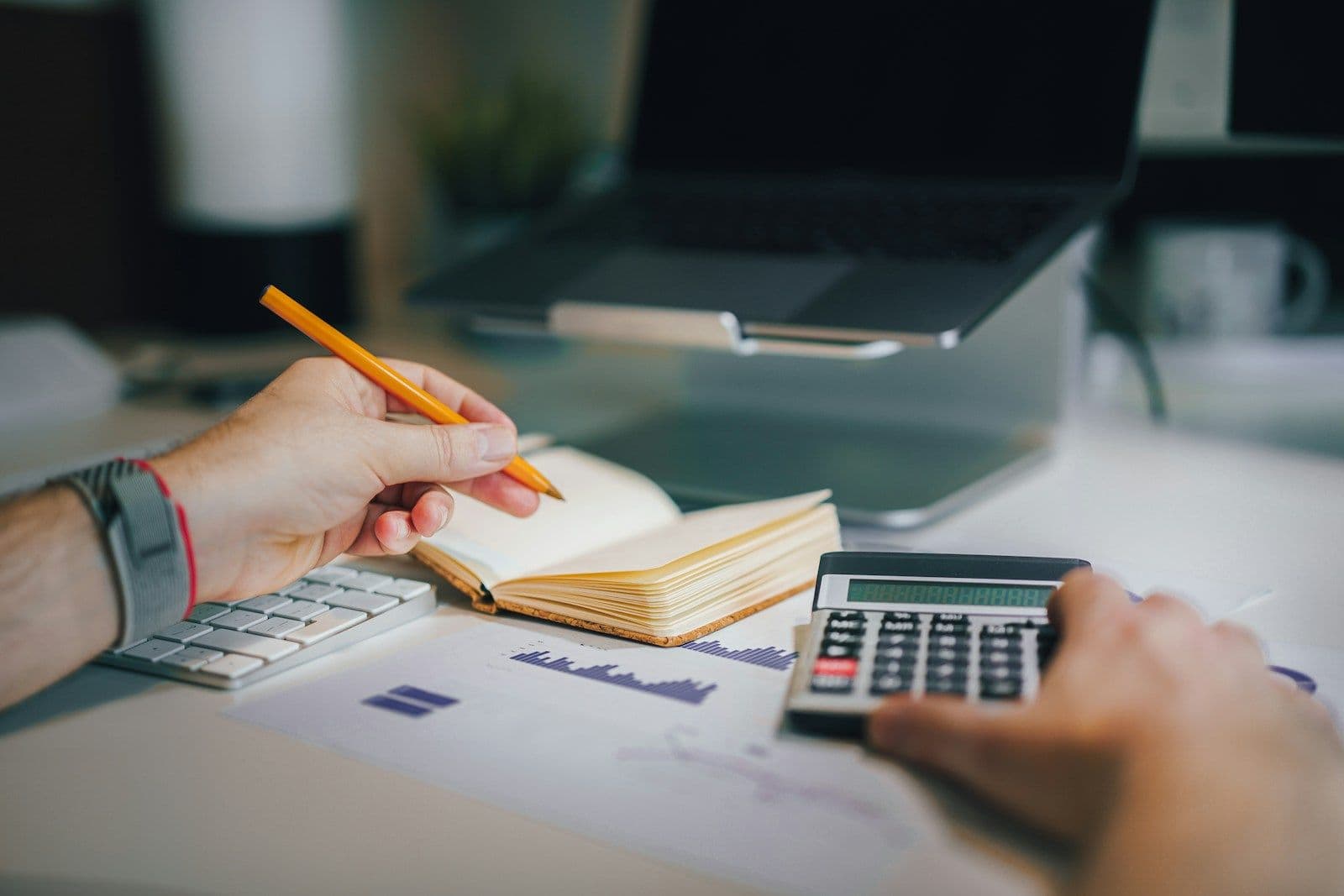 Person’s hands holding a pencil over a notebook while using a calculator beside printed financial charts on a desk.