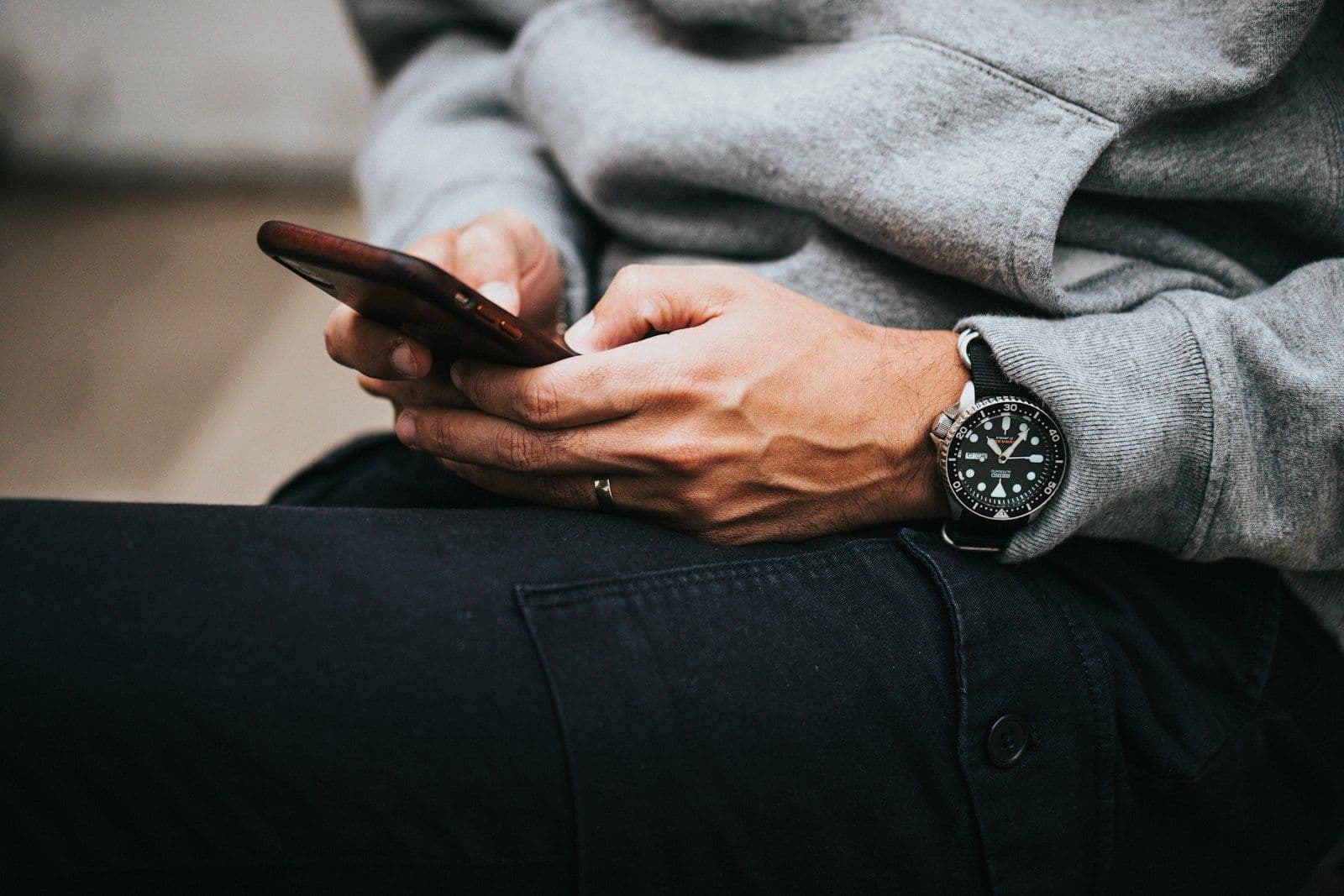 Close-up of a person in a gray hoodie and black pants holding a smartphone with both hands, wearing a silver wristwatch with a dark dial.
