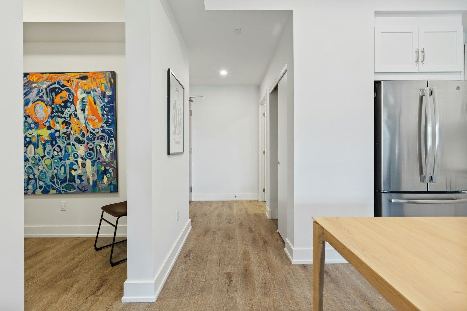 Bright apartment hallway with wood-look flooring, white walls, colorful abstract painting, and a stainless steel refrigerator