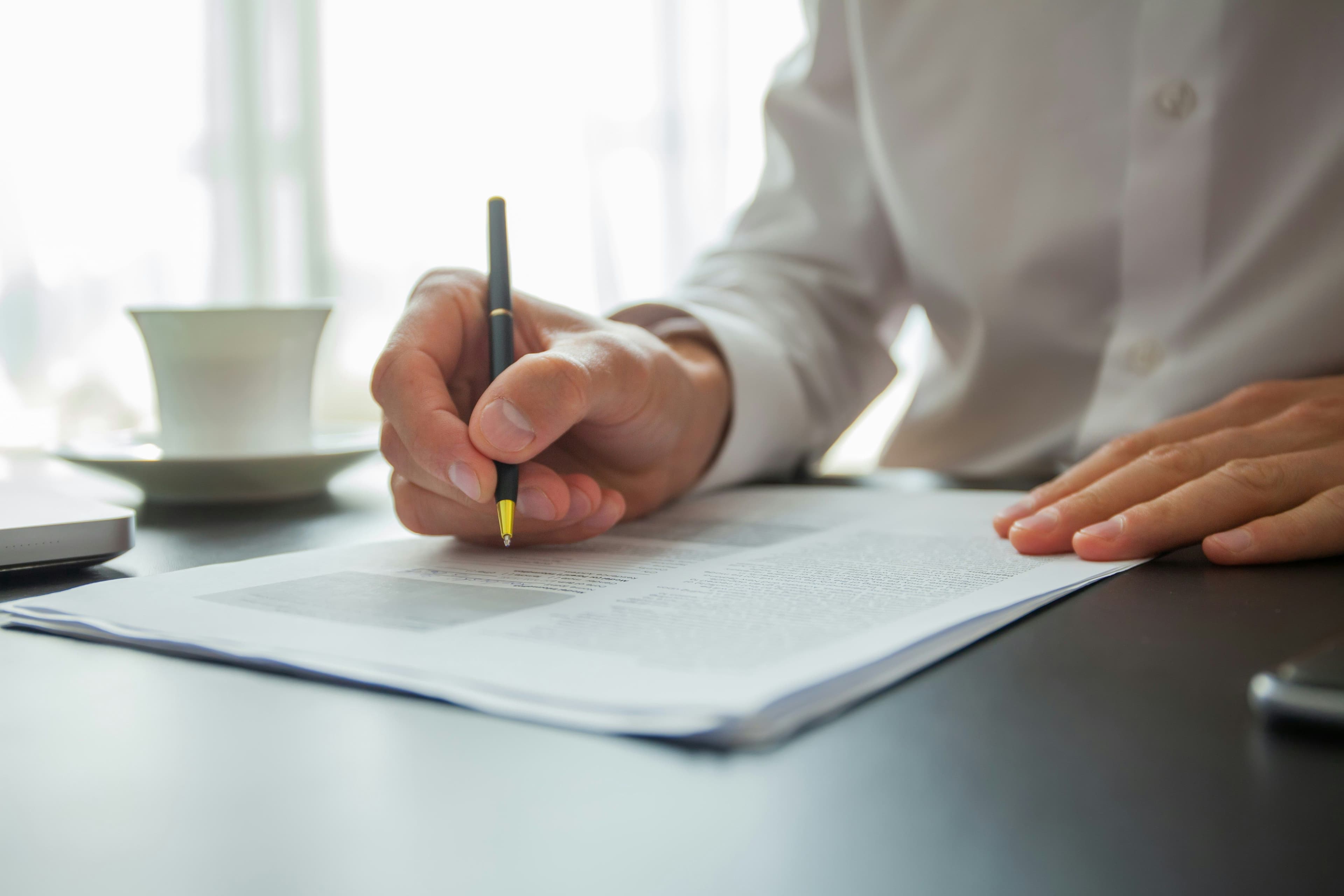 Hand in a white shirt writing on a document with a pen, with a coffee cup in soft window light.