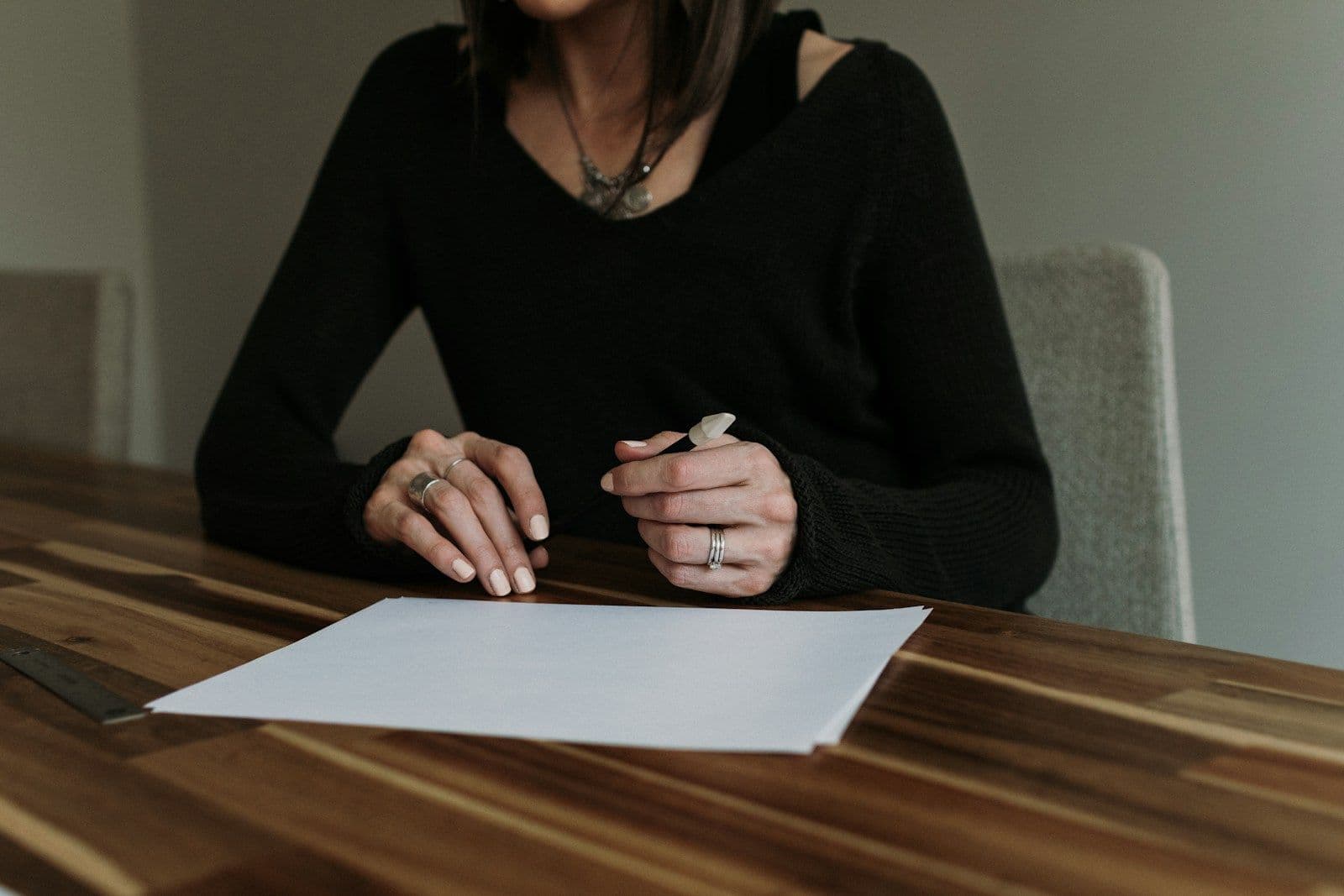 A woman in a black sweater sits at a wooden table, holding a pen above a blank sheet of paper, with rings on her fingers.