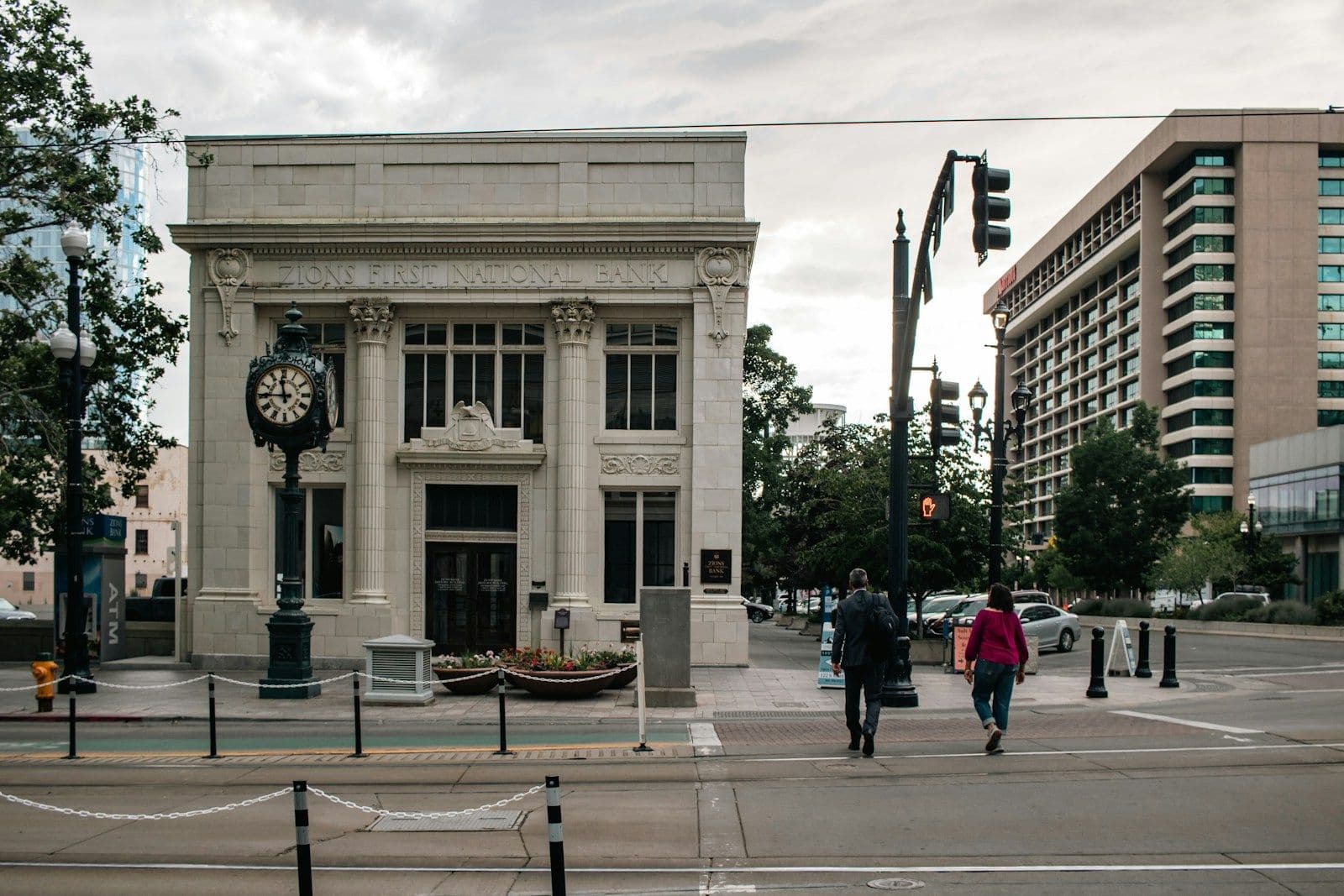 A pale stone bank building with a large ornate clock on a corner, with two pedestrians crossing a city street on a cloudy day
