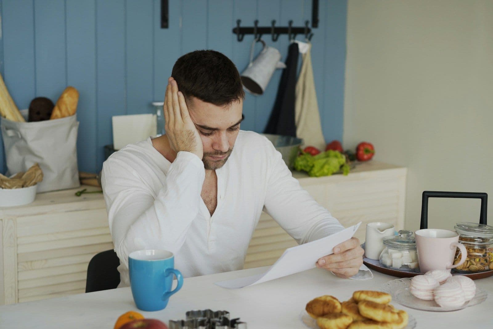 A man sits at a kitchen table holding a paper, resting his head on his hand with a concerned, thoughtful expression.