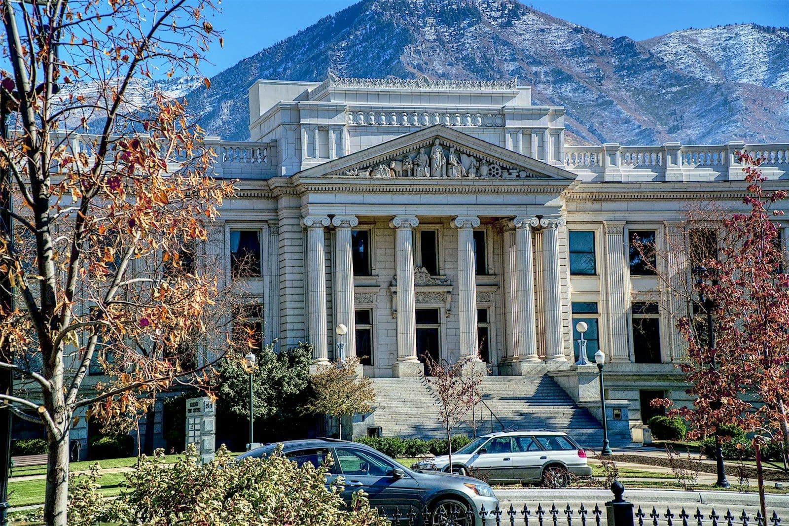 Neoclassical stone courthouse with tall columns and a pediment, set against snowy mountains, with parked cars and autumn trees in front.