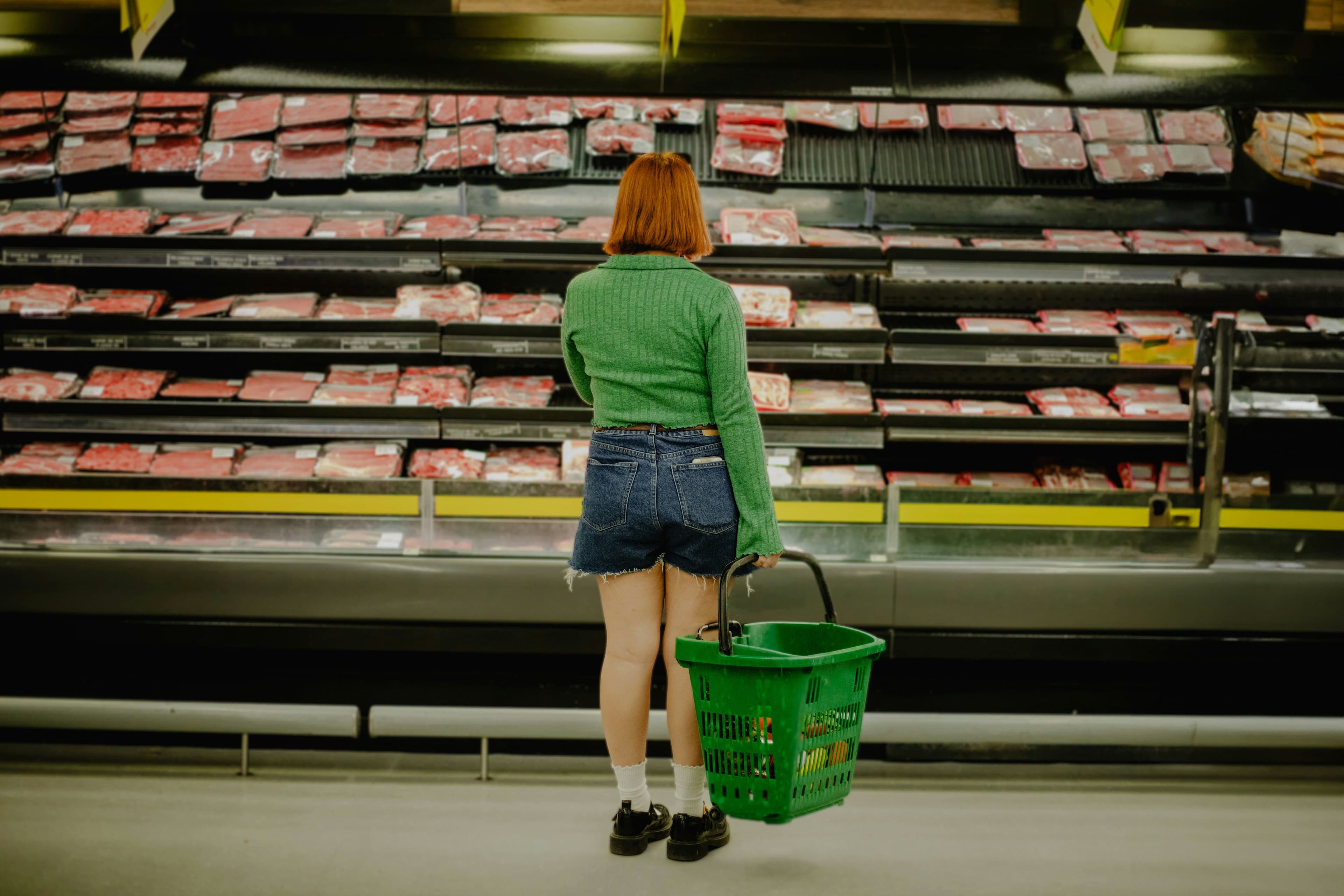 Woman in green sweater and denim shorts holding a green basket, standing in front of a supermarket meat case.