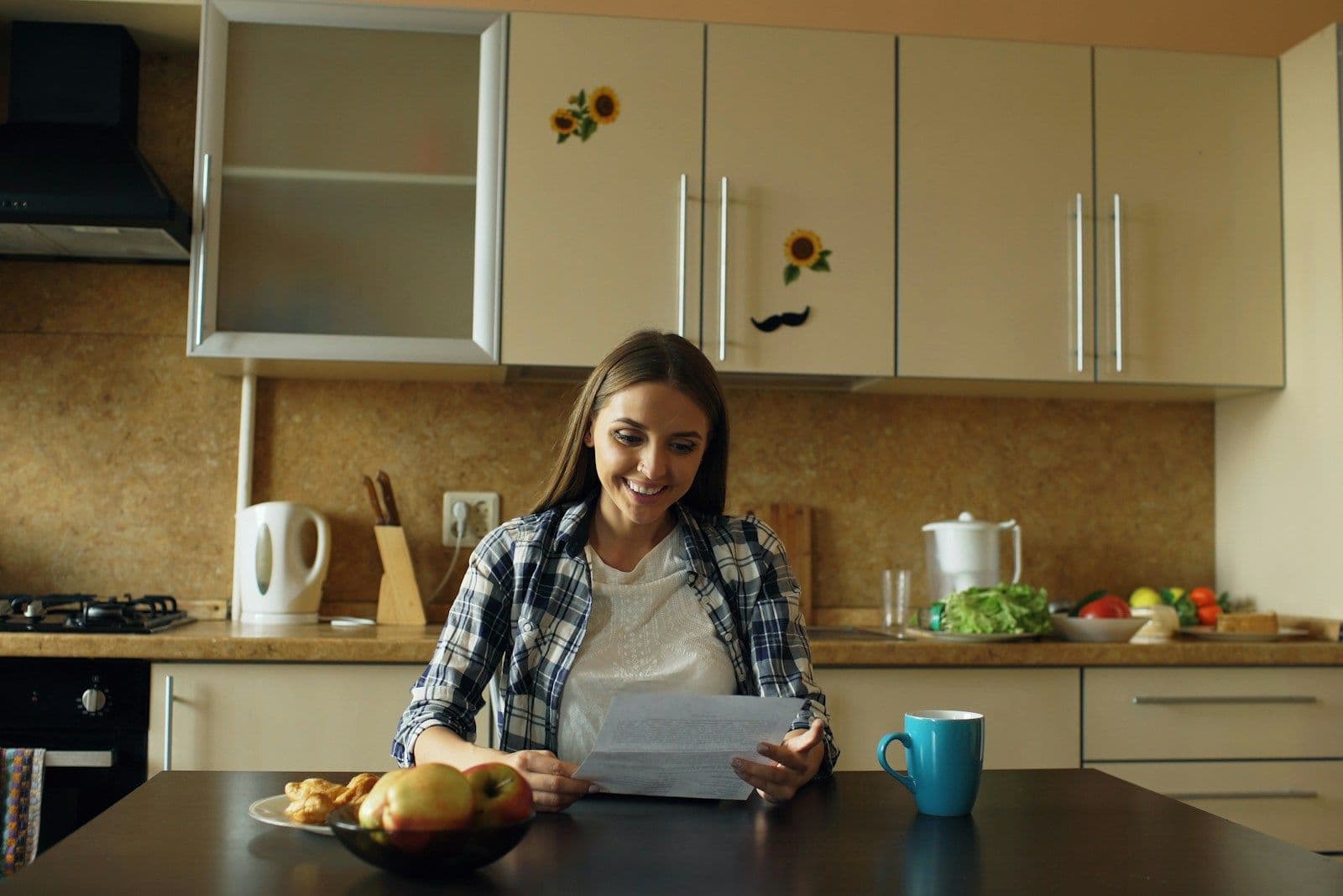 A woman sits at a kitchen table reading a paper document with a mug and a bowl of fruit nearby.