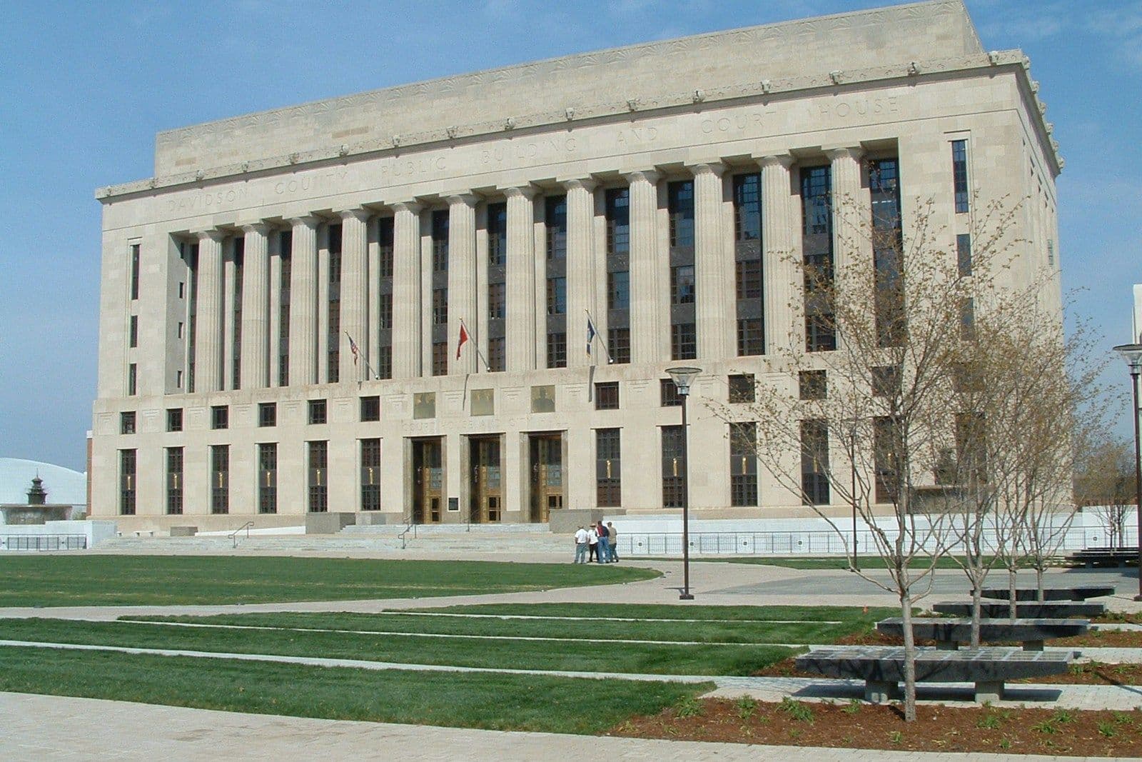 Large neoclassical stone government building with a row of tall columns, grassy lawn, and bare young trees in front on a clear day