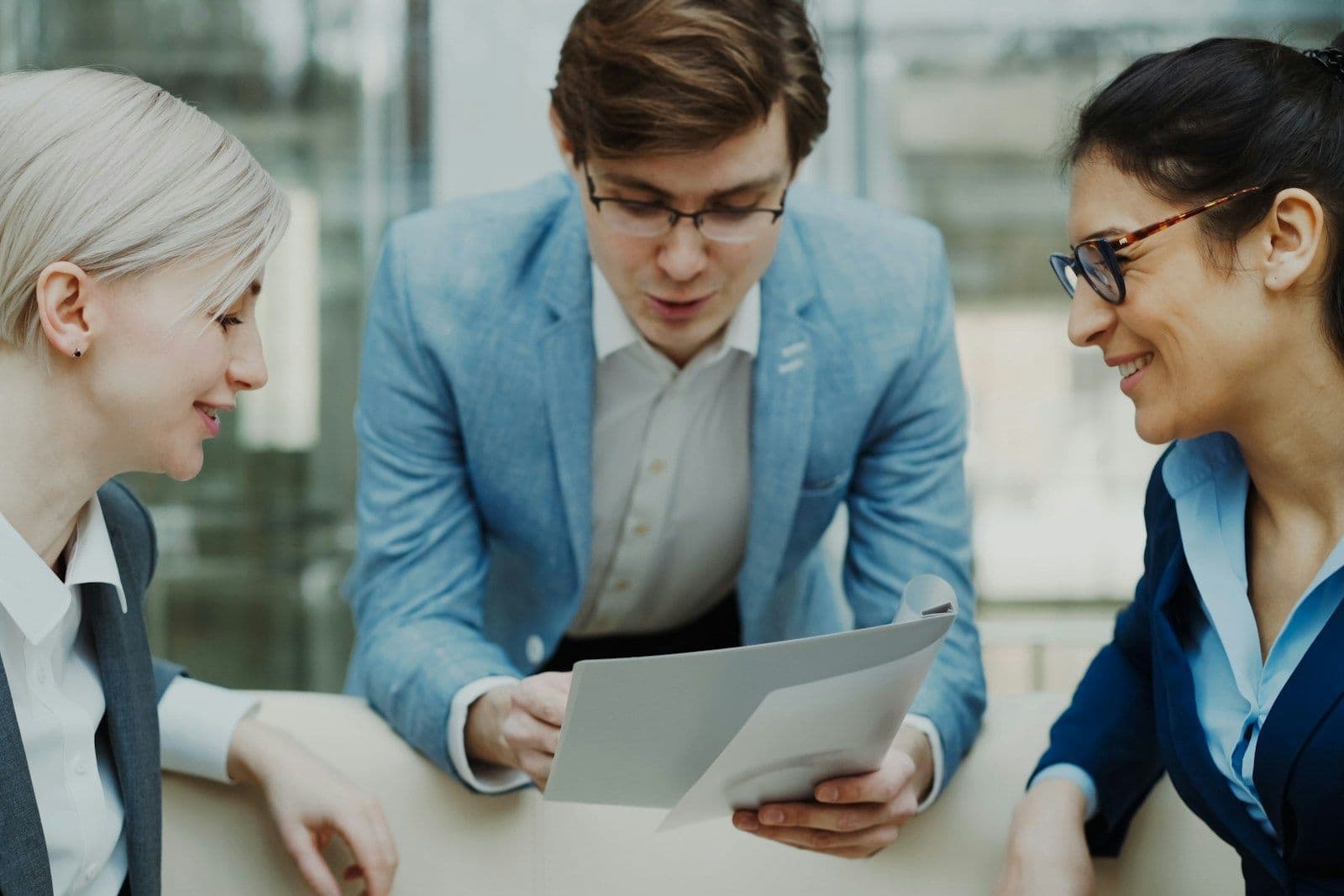 Three colleagues in business attire review a document together at a table, smiling and engaged in discussion.
