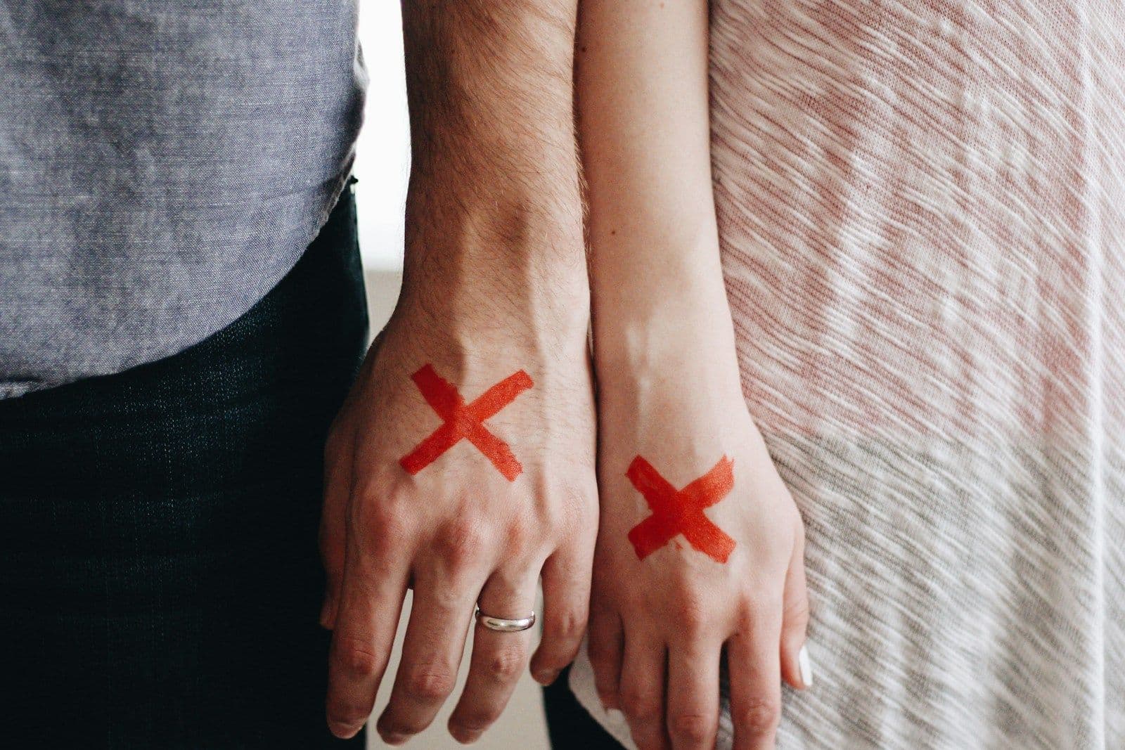 Two people’s hands held together, each marked with a bold red X on the back of the hand, one wearing a silver ring