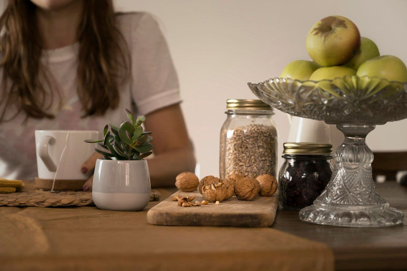 A person seated at a wooden kitchen table with a mug, a small potted plant, walnuts, jars, and a bowl of apples.