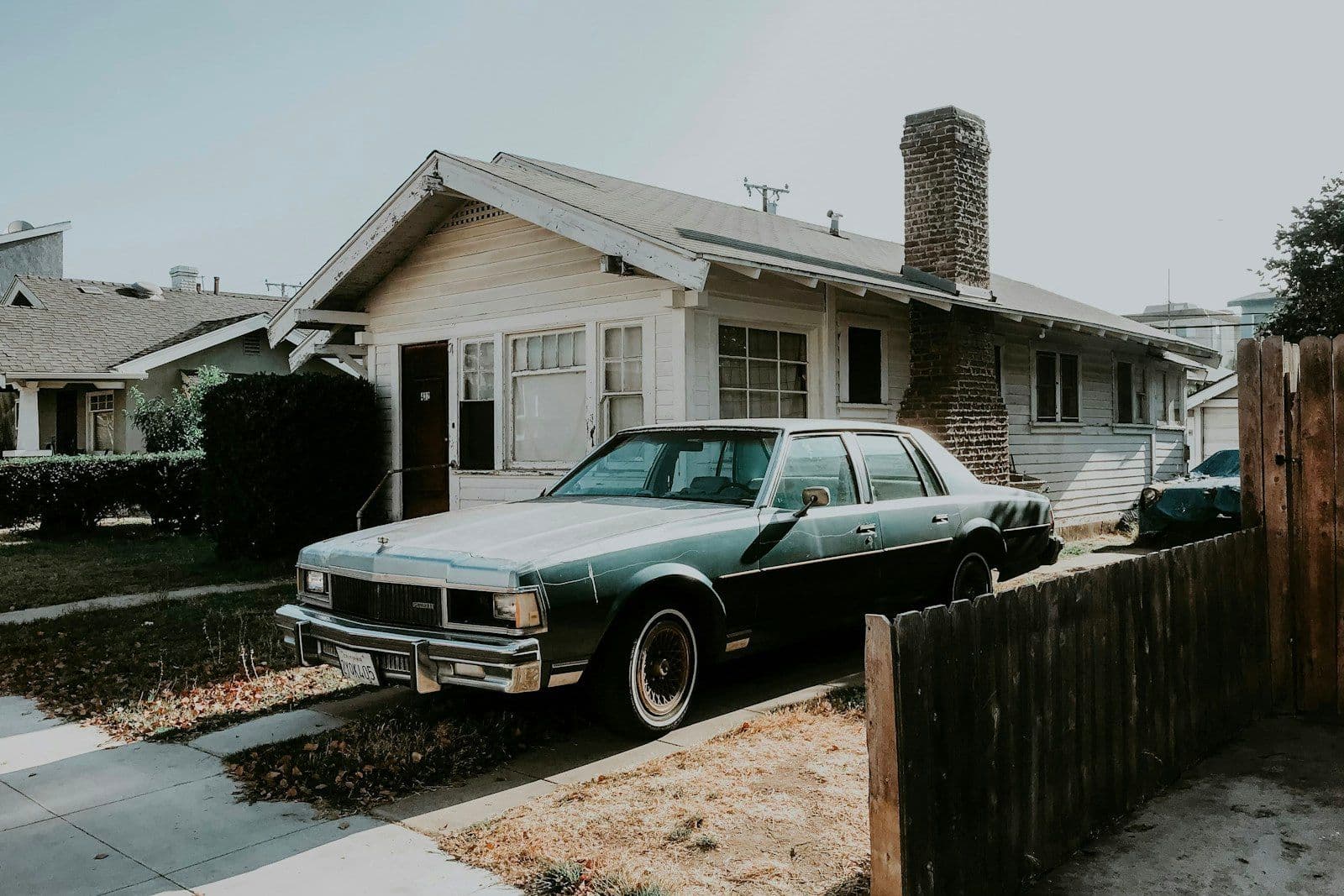 A dark green vintage sedan parked in the driveway of a single-story bungalow with a brick chimney and wooden fence.