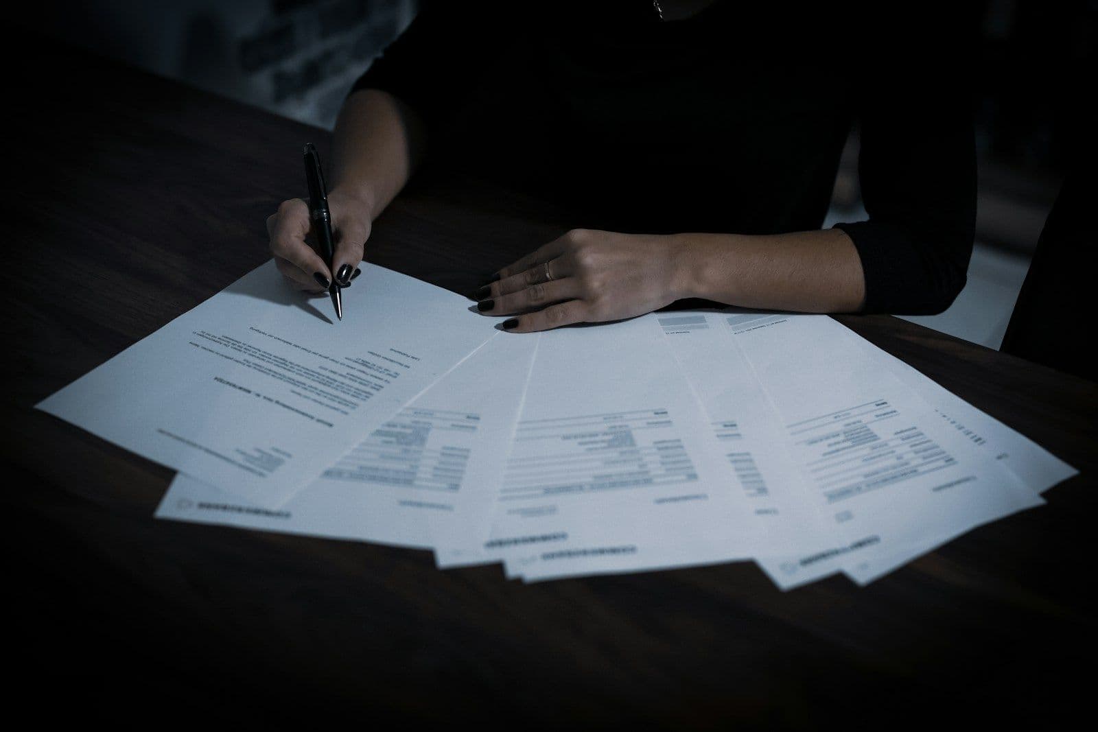 A person’s hand writing with a pen on paper documents in dim lighting, viewed from above.