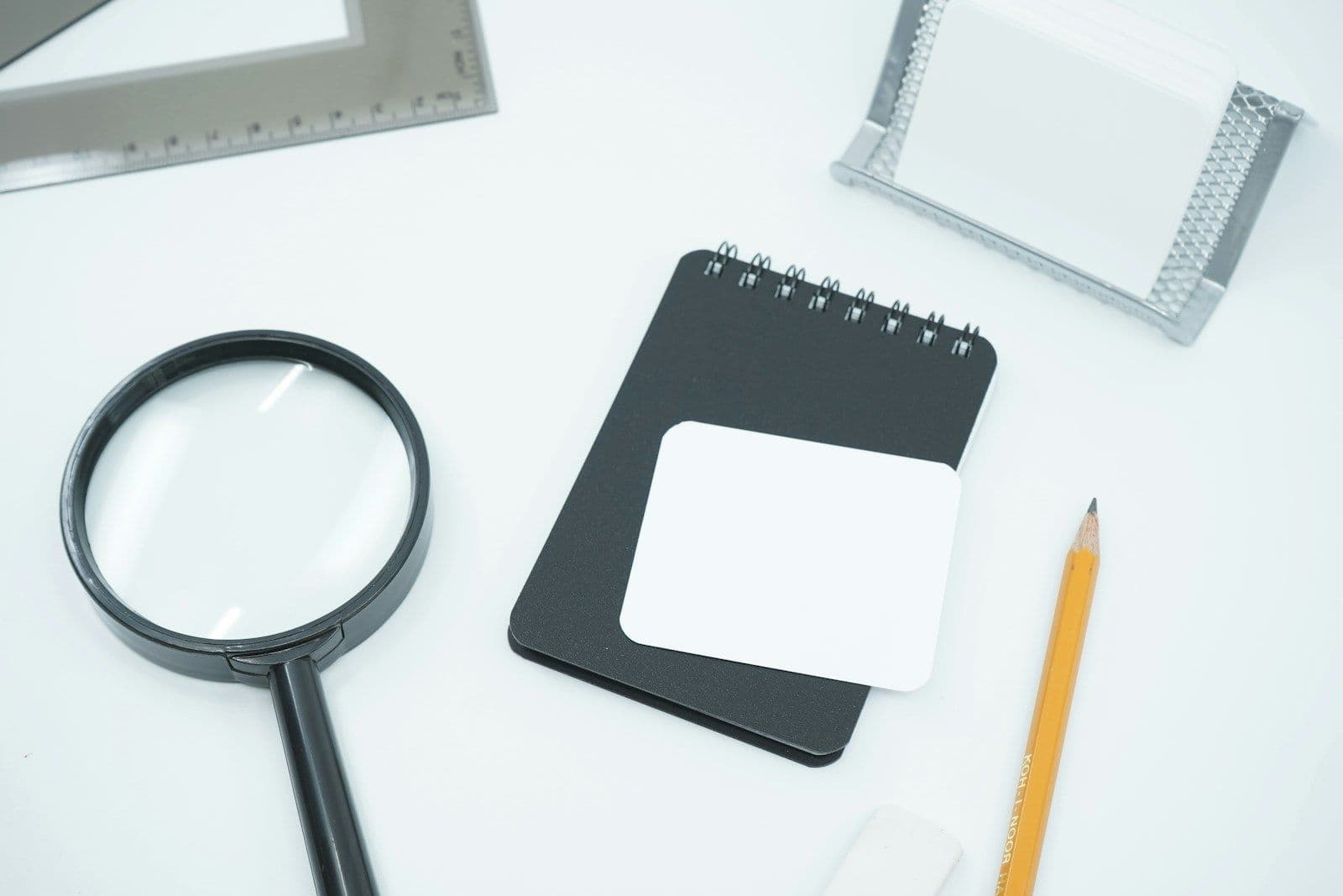 A magnifying glass, notebook and pencil on a white desk