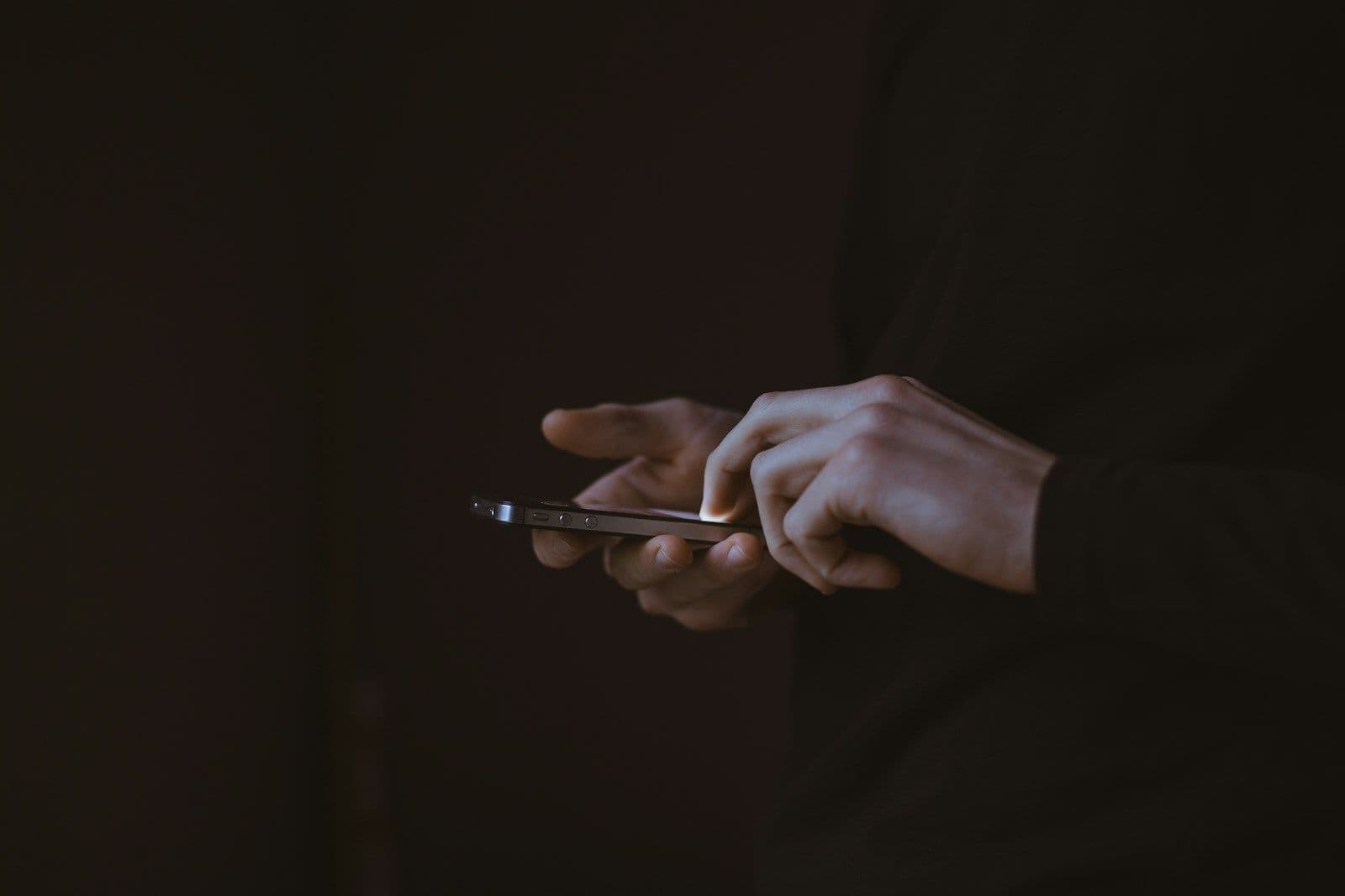 Close-up of hands holding a smartphone in a dark setting, with the screen lit faintly as the person types or scrolls.