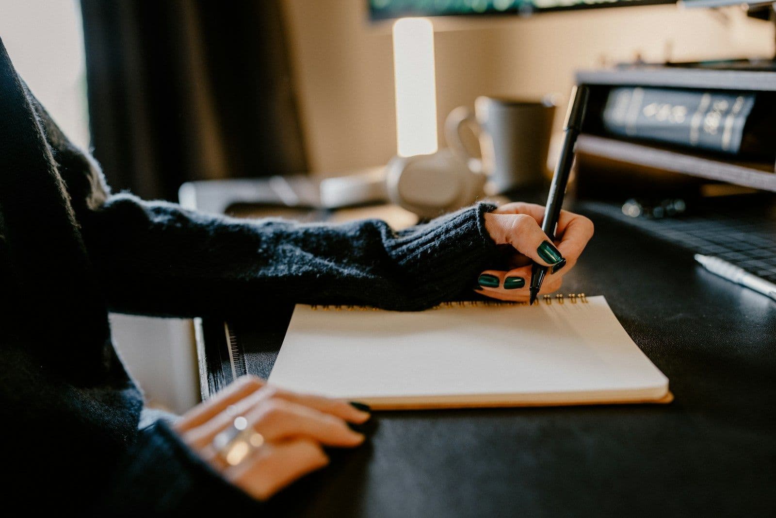 A person in a dark knit sweater writes with a pen in a spiral notebook on a dark desk, with a mug and monitor in the background