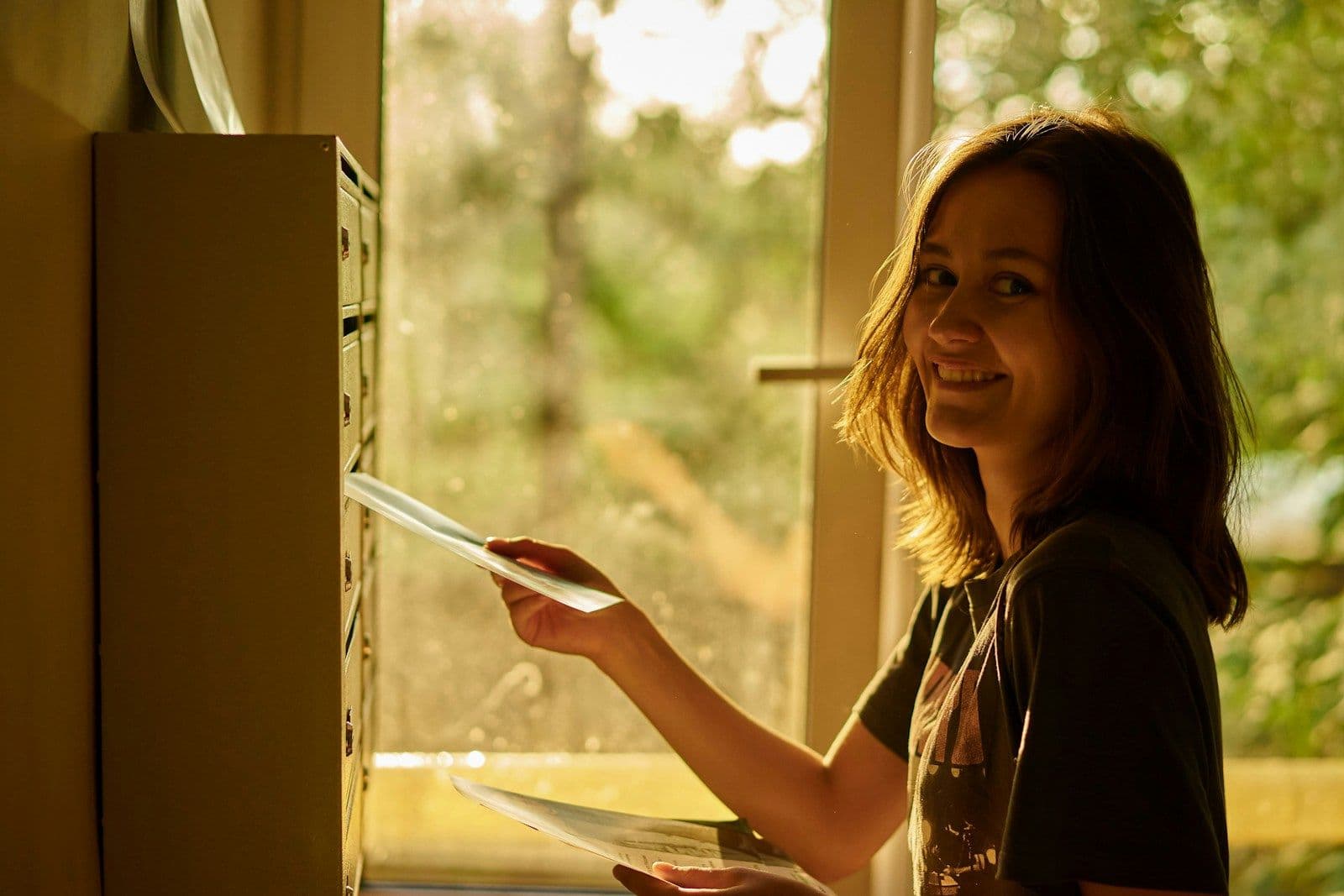 Smiling woman in warm window light collecting letters from a row of apartment mailboxes.