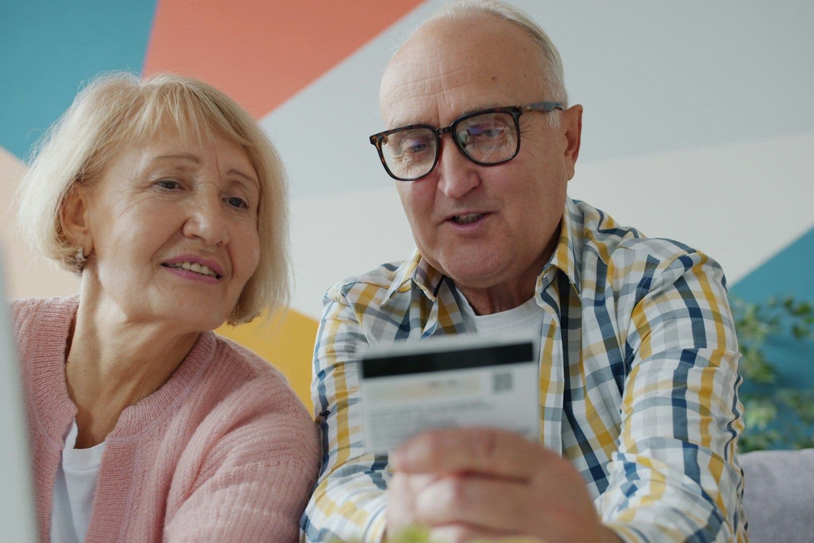 An older couple sits side by side looking at a laptop while the man holds up a credit card and reviews it together with the woman, who smiles beside him.