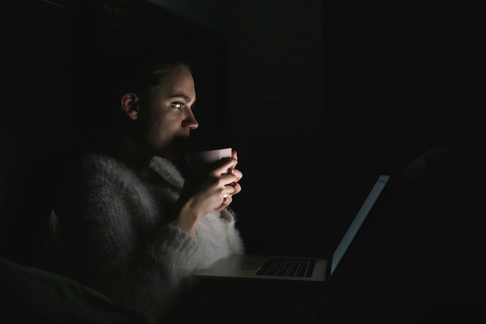 A woman in a dark room holding a mug, lit by her laptop screen