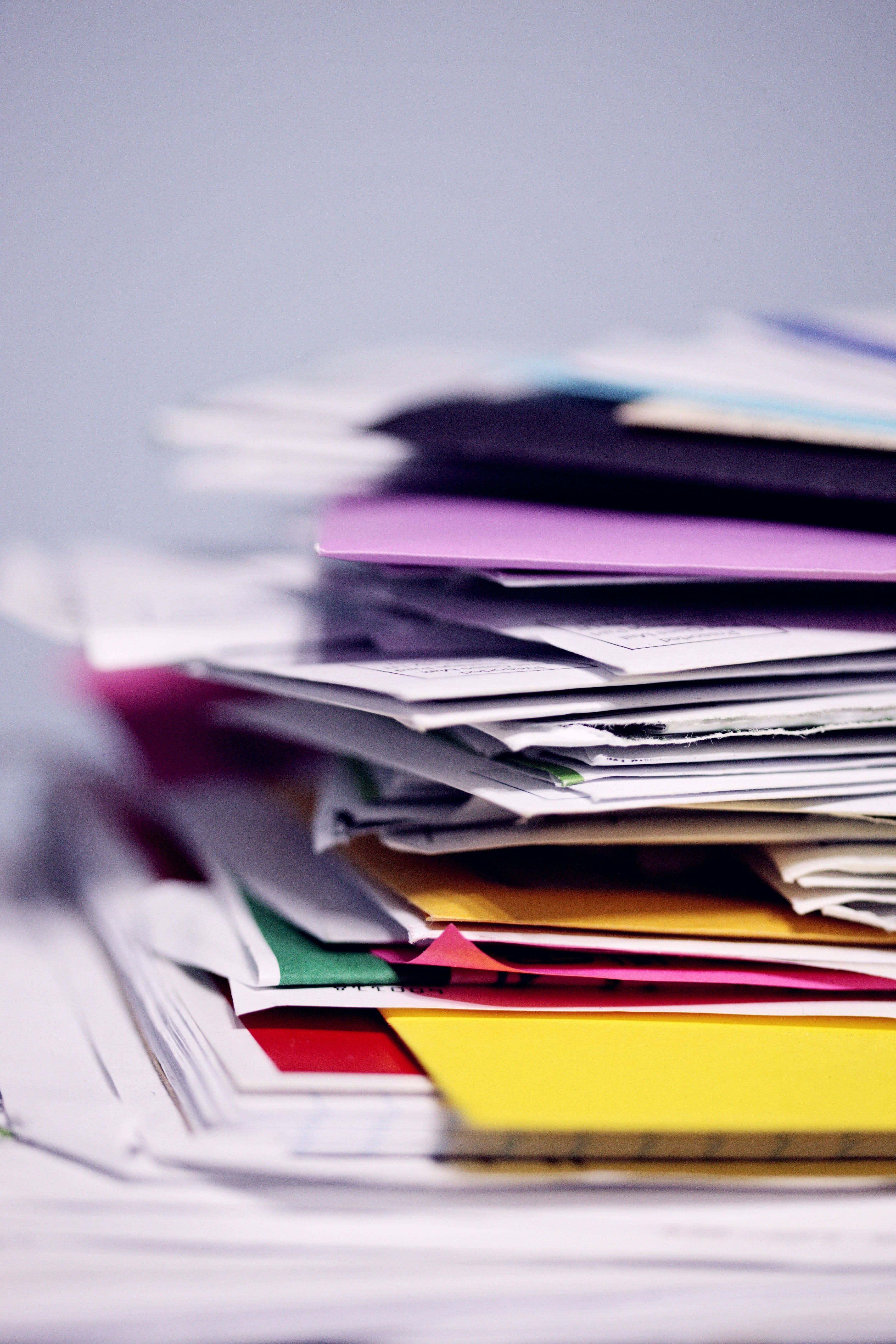 Tall stack of mixed papers, envelopes, and colored folders against a soft gray background.