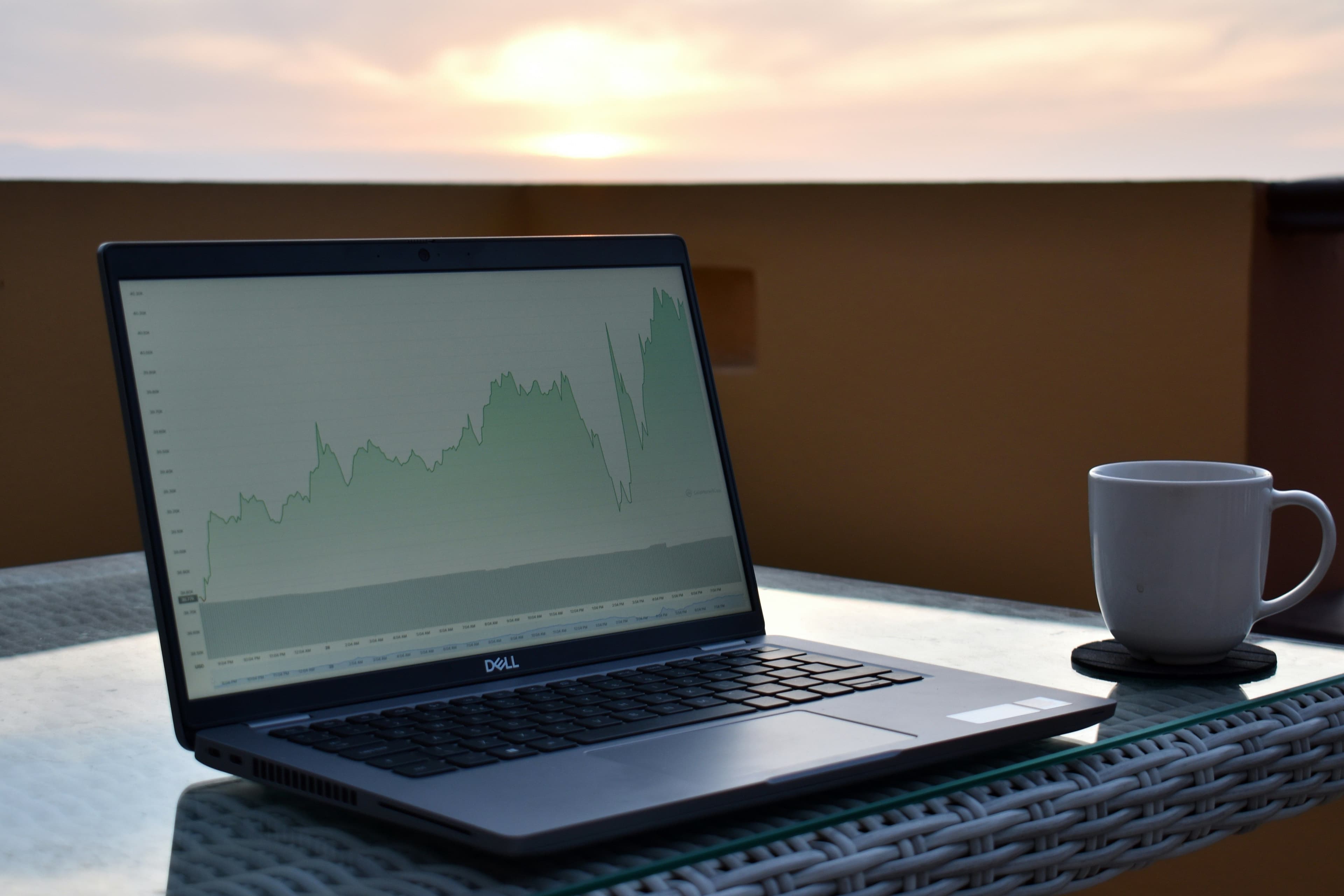 Open Dell laptop on a glass patio table displaying a rising green line chart, with a white coffee mug beside it at sunset.