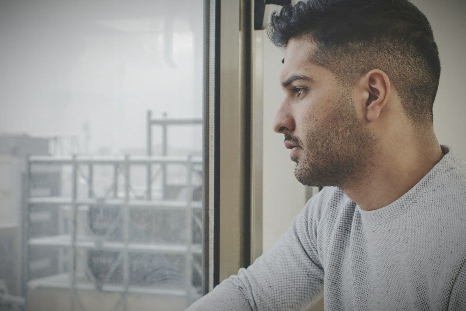 Profile of a bearded man in a gray sweater gazing thoughtfully out a window with a blurred urban scene behind him.