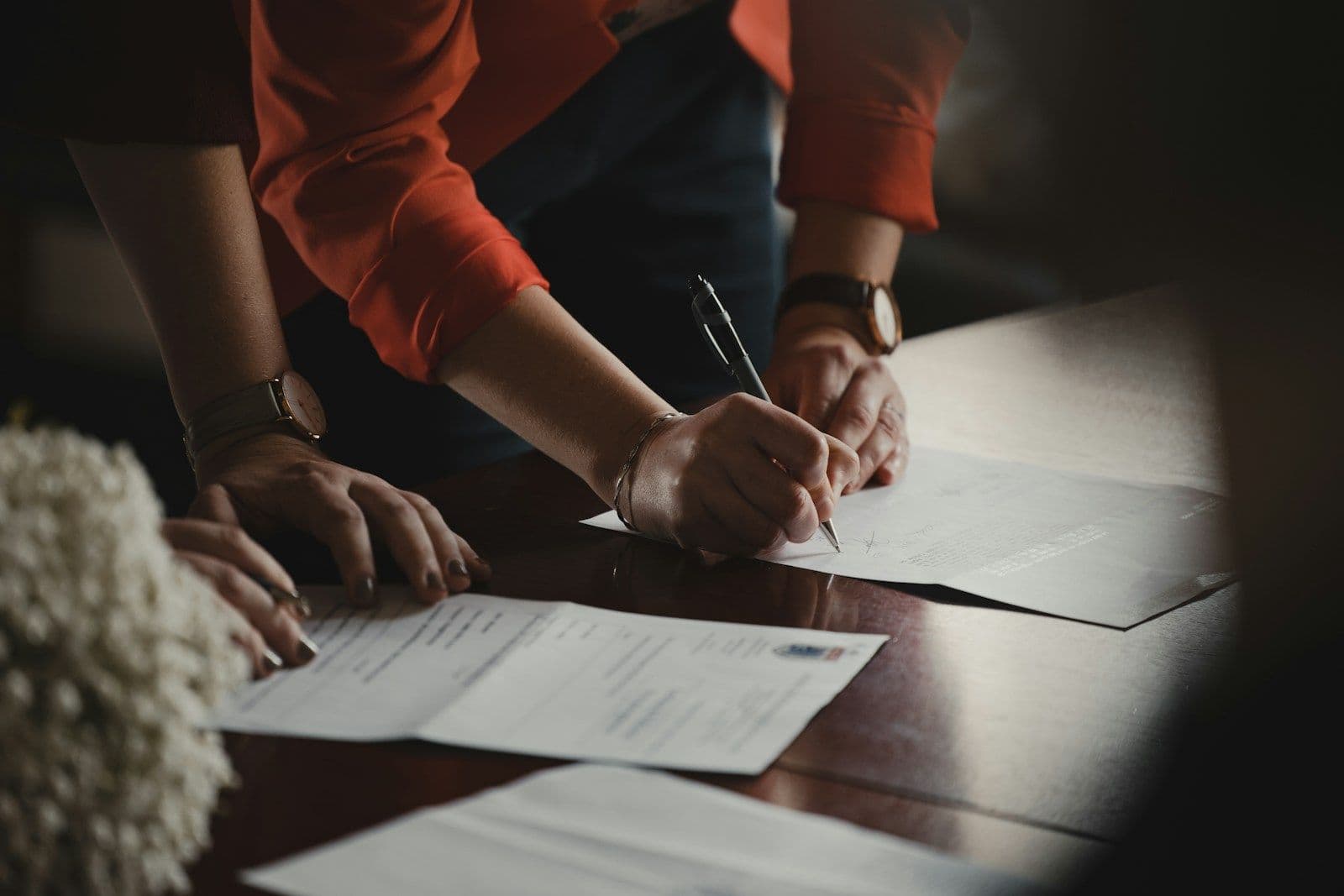 Close-up of two people’s hands over paperwork on a wooden desk, one hand signing with a pen
