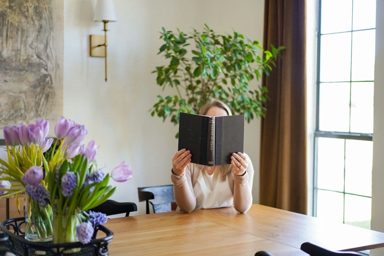 A woman sits at a wooden table reading a dark book, with purple tulips in the foreground and a window behind her.