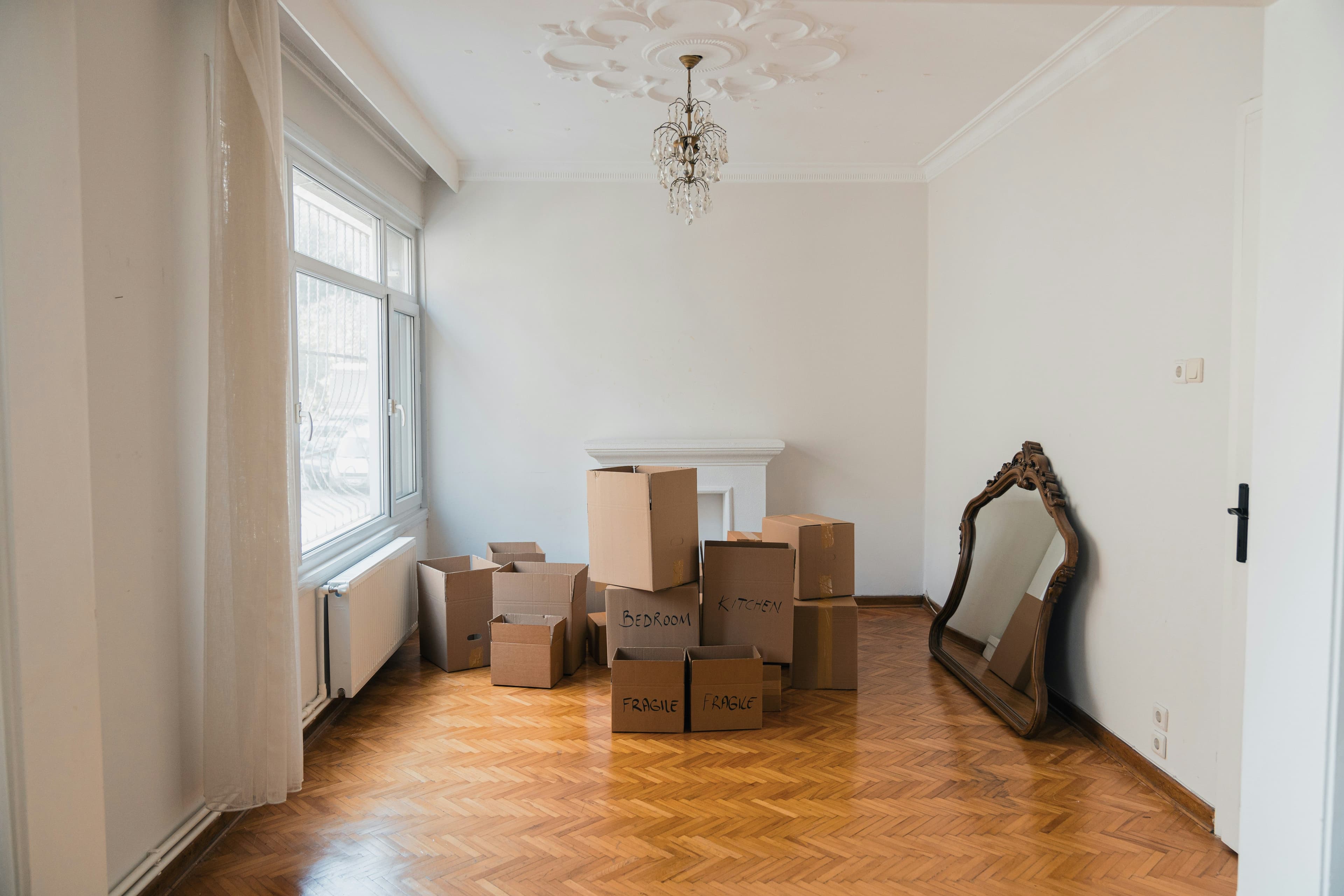 Empty room with wood floors, stacked moving boxes labeled Bedroom and Kitchen, and an ornate mirror against the wall.