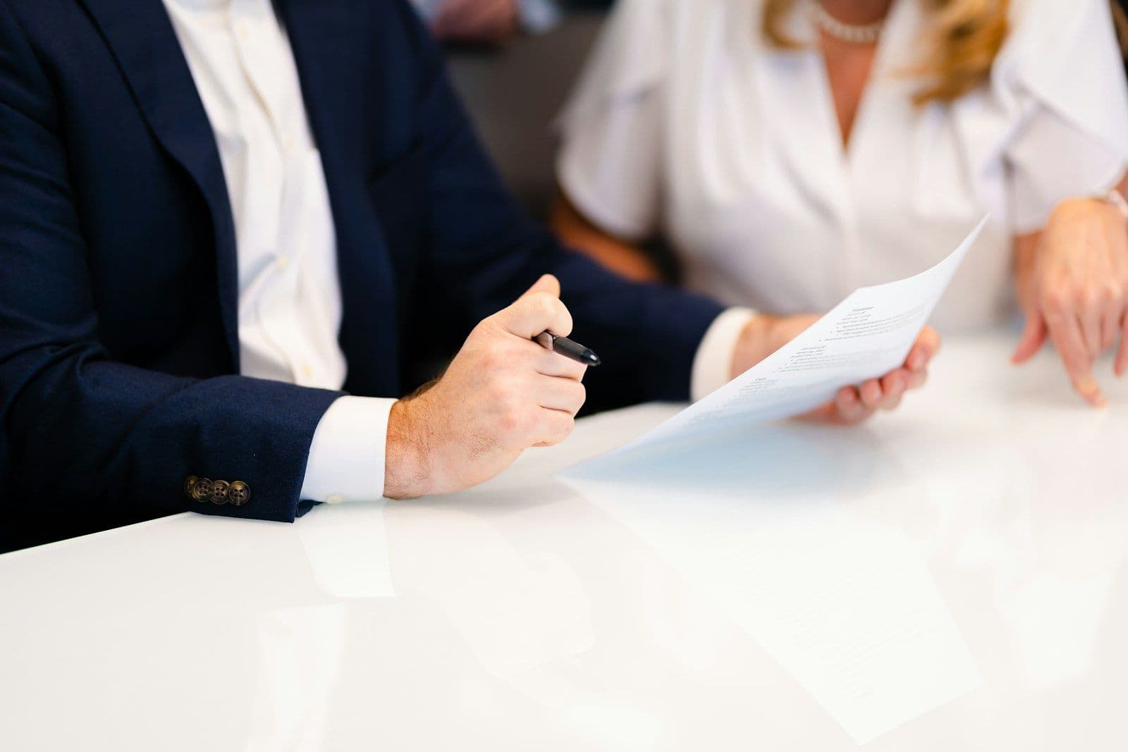 A man in a navy suit holds a pen and reviews a document at a white table, with a woman in a white blouse beside him reading the same paper.