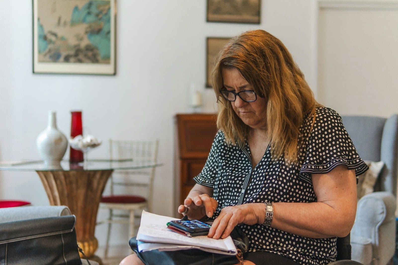 A woman with glasses and shoulder-length hair sits in a living room using a calculator while reviewing paper documents on her lap.