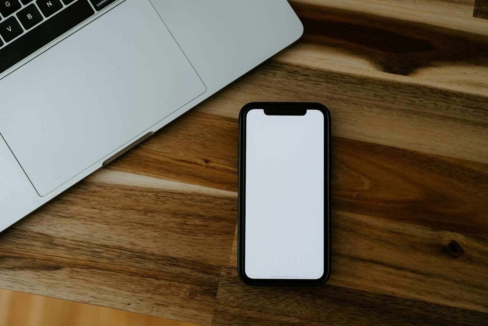 Overhead view of a silver laptop and a black smartphone with a blank white screen on a wooden desk surface.