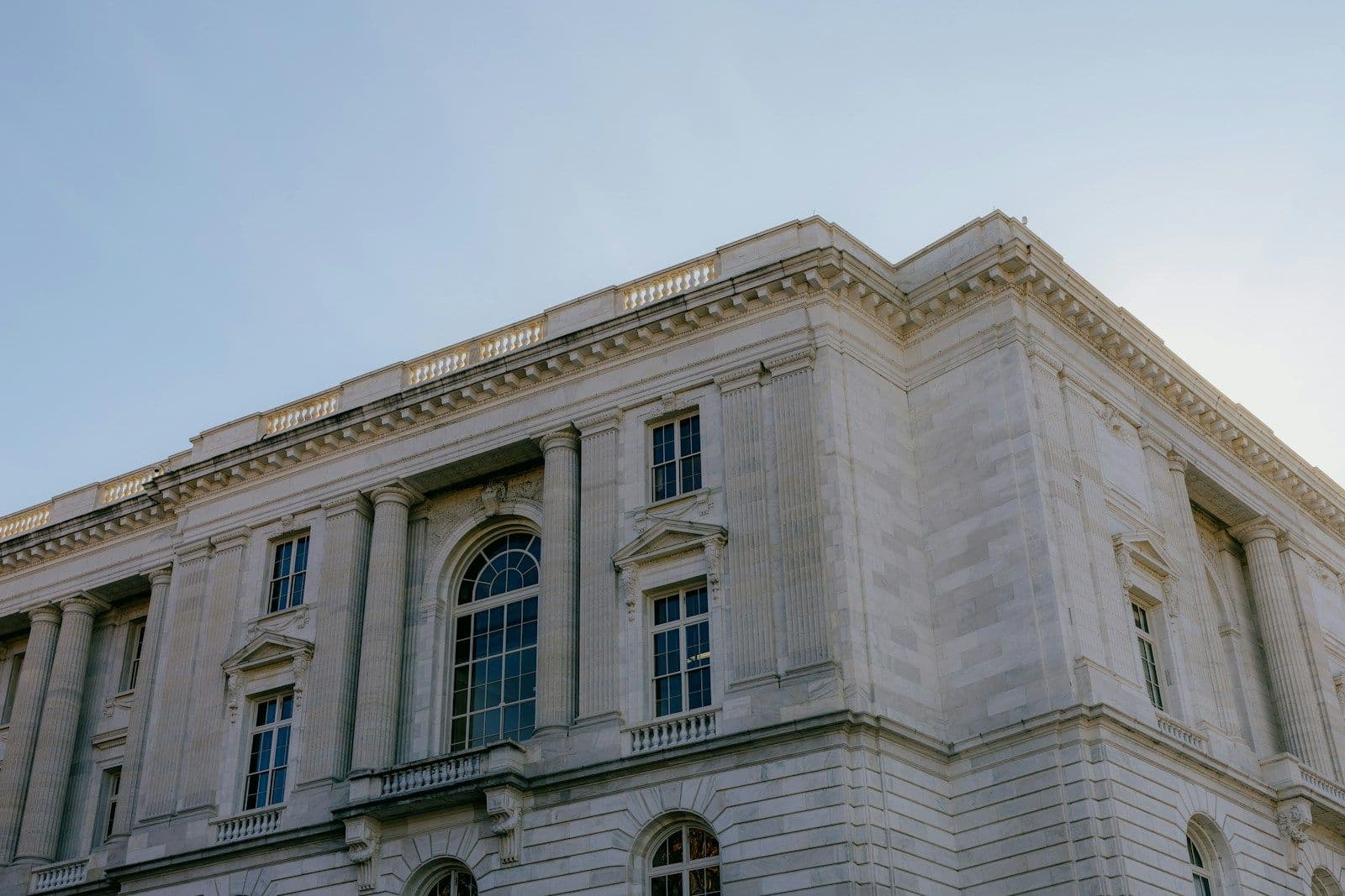 Looking up at a large white stone government-style building with tall arched windows, columns, and a decorative cornice under a pale blue sky