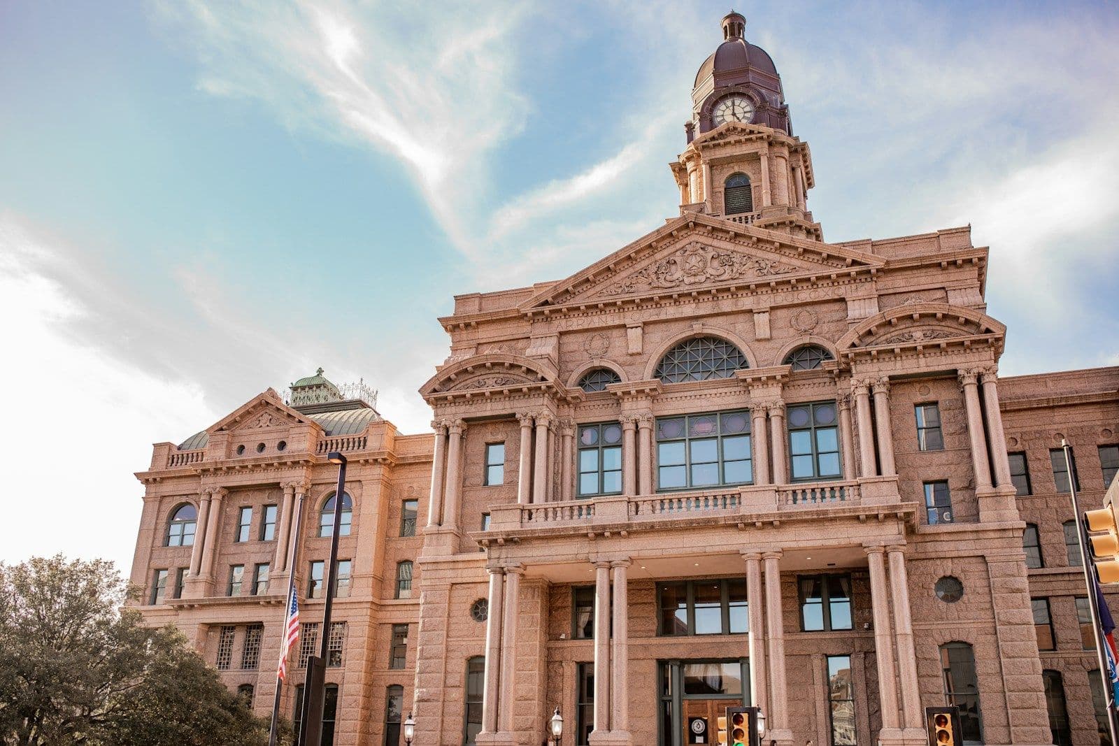 Ornate pink sandstone county courthouse with a clock tower, columns, and American flags under a partly cloudy blue sky
