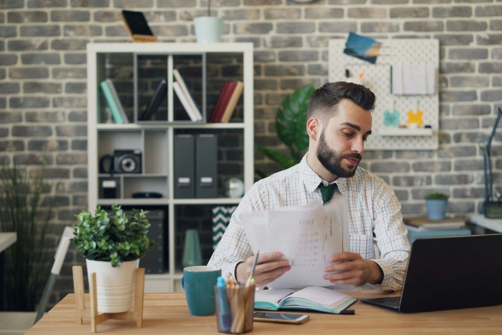 A bearded man in a striped shirt and tie reviews printed papers at a wooden desk with an open laptop, notebook, and coffee mug in a home office.
