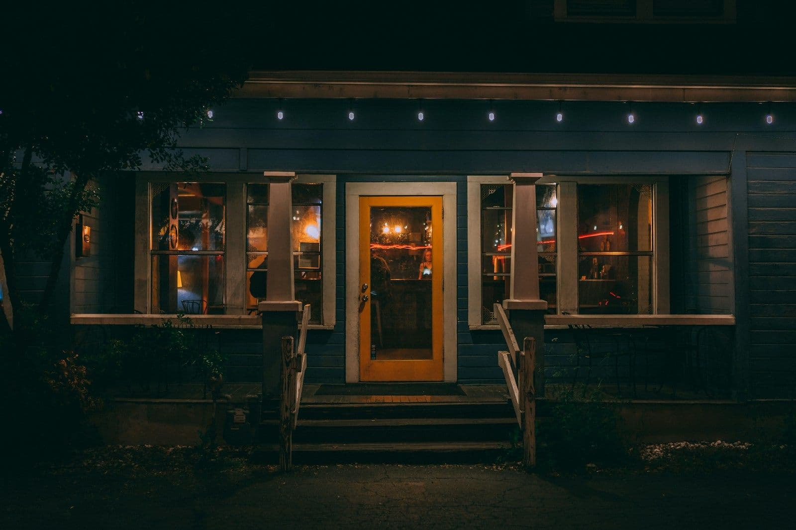 A small blue house at night with warm yellow light glowing from its front door and windows, with string lights along the roofline and a few steps leading up to the porch.