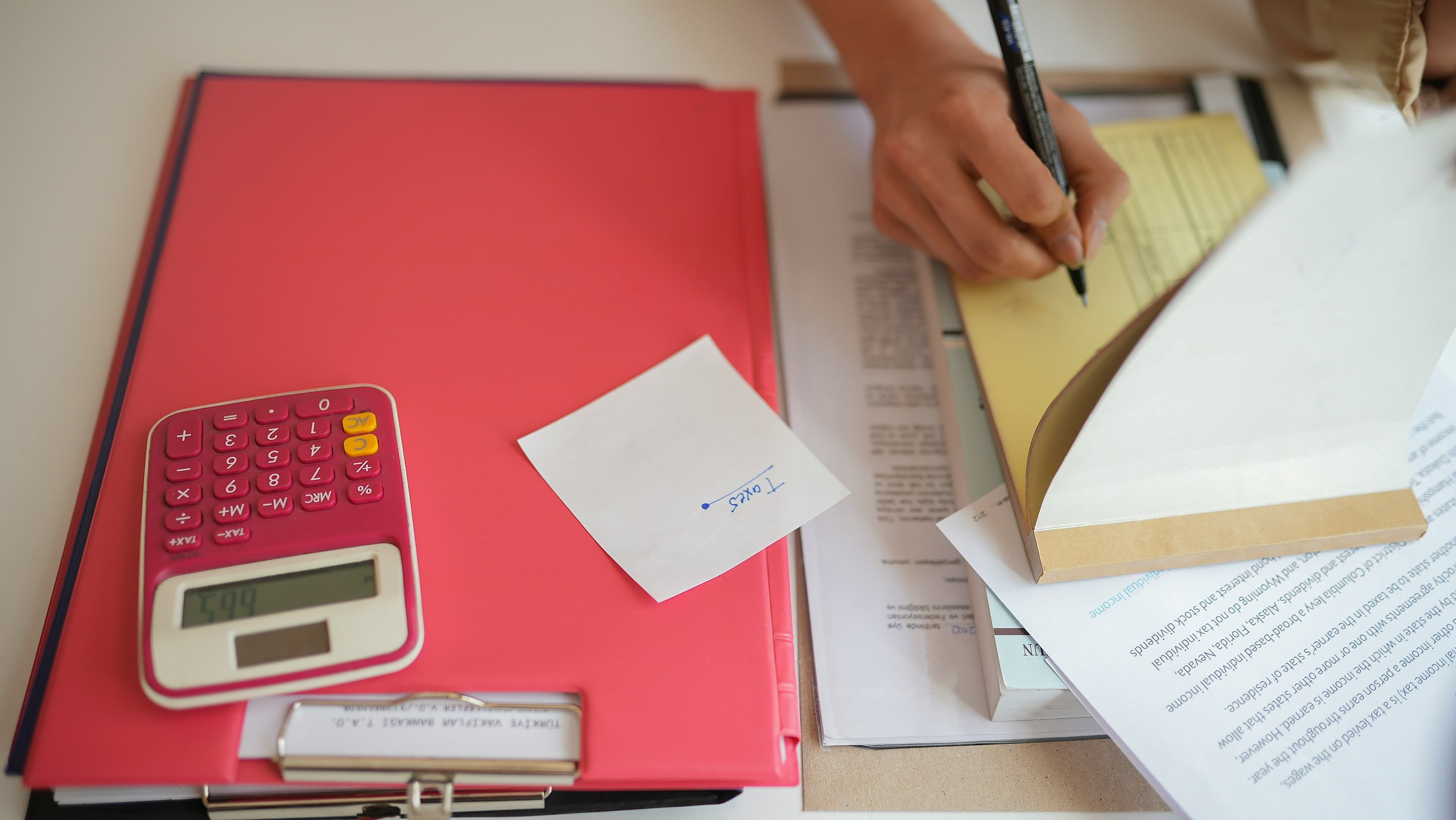 Hand writing on paperwork beside a red calculator showing 655 and a stack of financial documents.