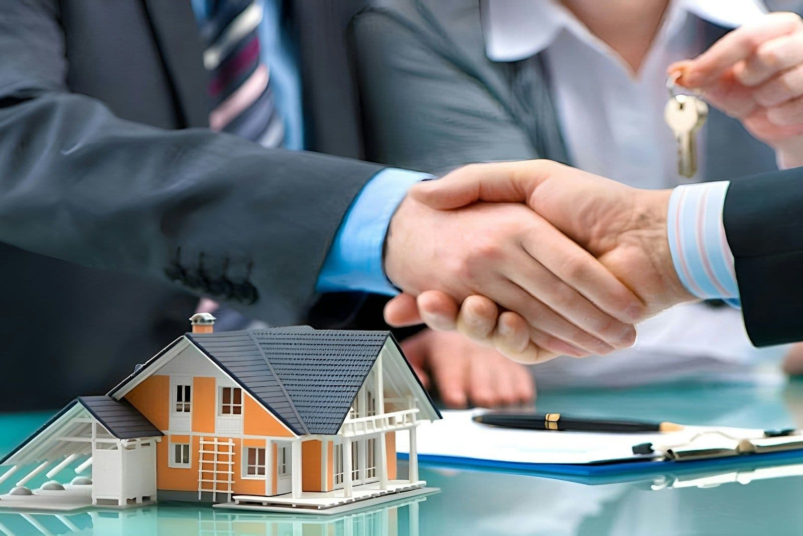 Two people shaking hands across a glass table beside a model house, a set of keys, and a signed document, suggesting a home purchase deal