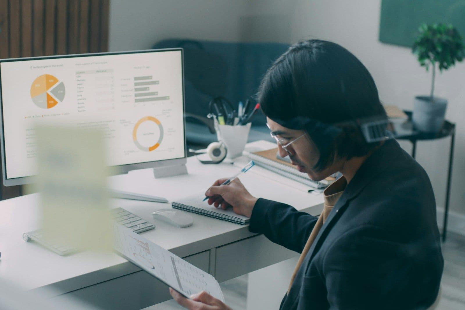 A person with a headset works at a desk in front of a monitor showing pie and donut charts, with a notepad and a potted plant nearby.