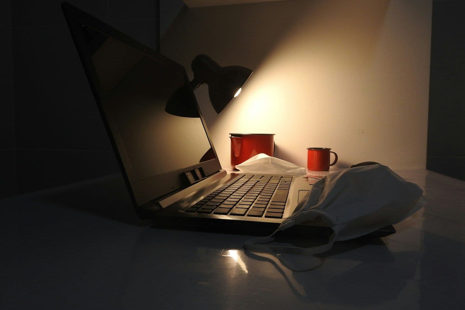 A dimly lit open laptop on a desk beside a desk lamp, a red mug, and a folded white face mask, with a warm glow against a dark background