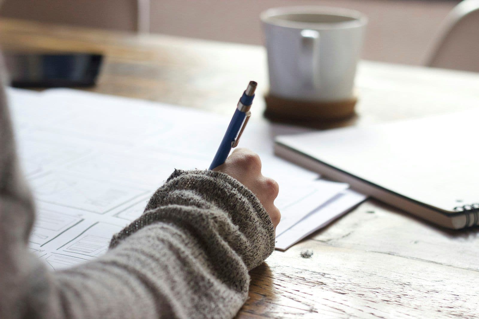 A person’s arm in a gray knit sweater writing with a blue pen on a white form at a wooden table, with a white mug and a closed notebook nearby.