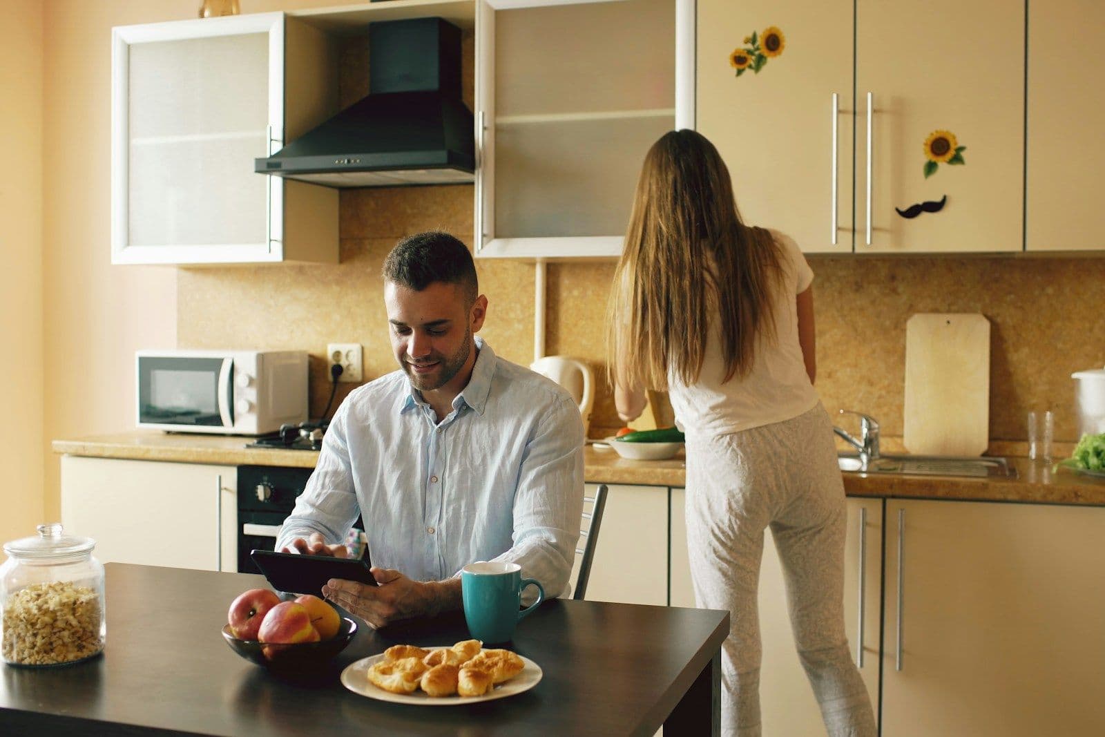 A man in a blue shirt sits at a kitchen table with pastries and fruit, looking at a device while a woman works at the counter.