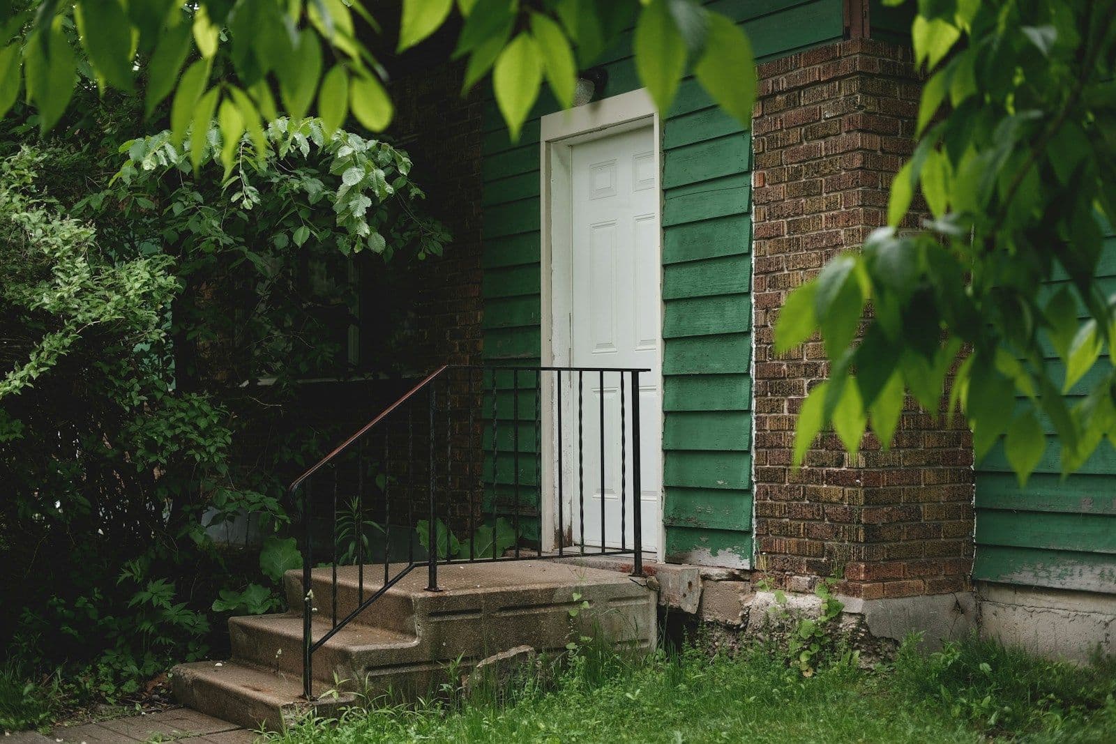 A white door at the top of worn stone steps on a green-sided house with a brick corner, framed by leafy trees and grass.