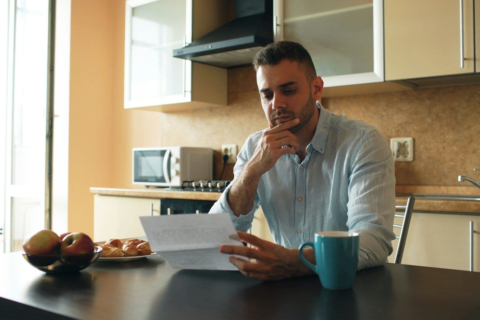 A man sits at a kitchen table with a hand to his chin, thoughtfully reviewing a paper beside a mug and a bowl of fruit.
