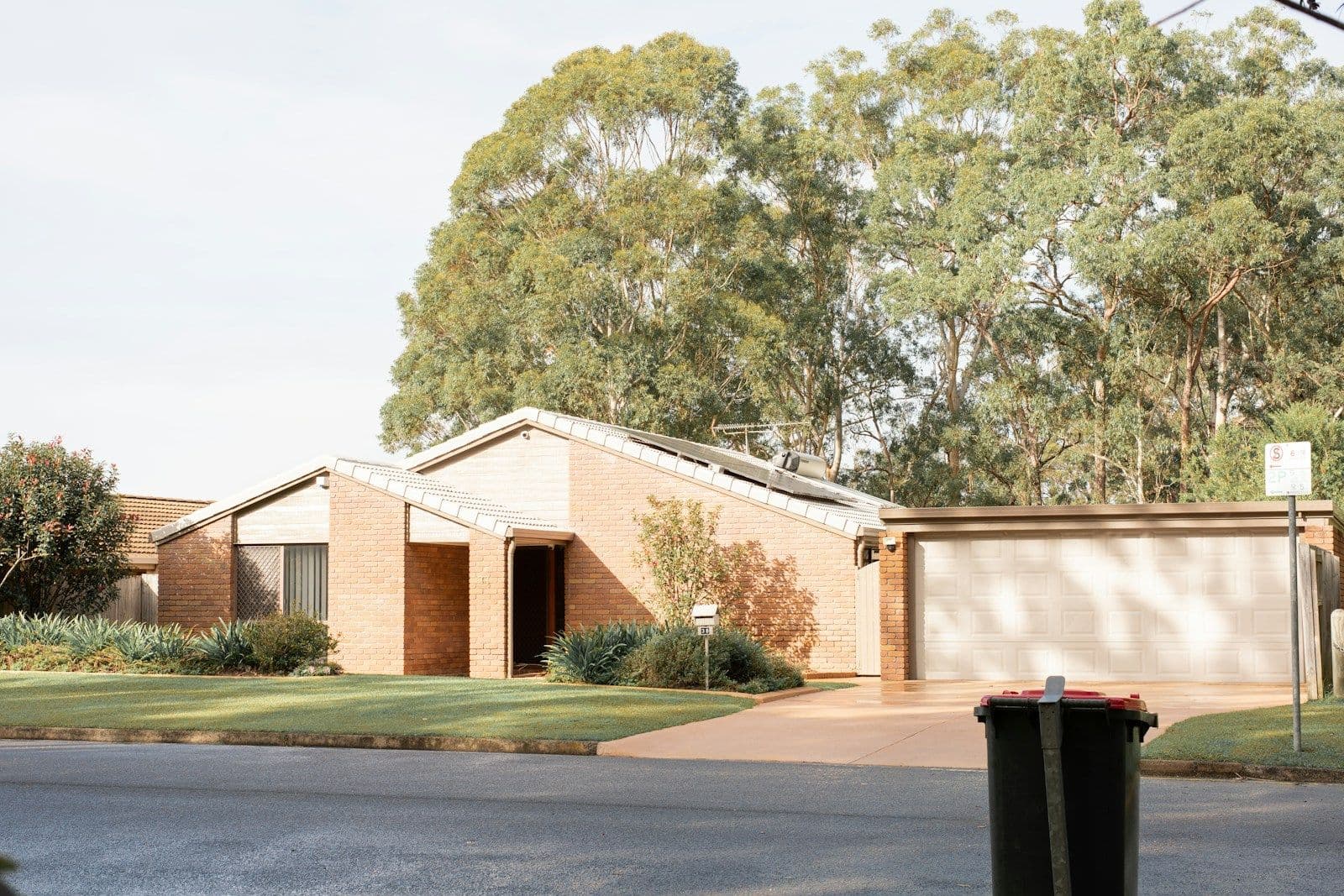 A brick suburban house with a garage and lawn on a quiet street, with a dark trash bin at the curb in soft daylight.