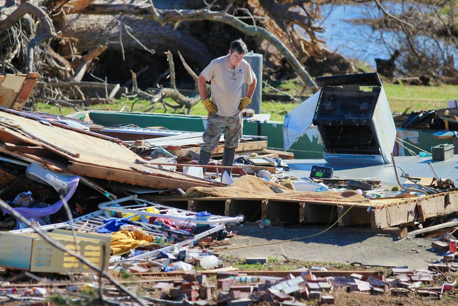 A man in work gloves stands among splintered wood and debris from a storm-damaged building beside a wooded waterway.