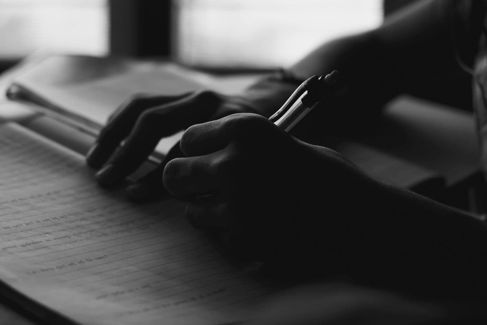 Black and white close-up of hands holding a pen over a document with printed text, suggesting filling out a form or writing a letter