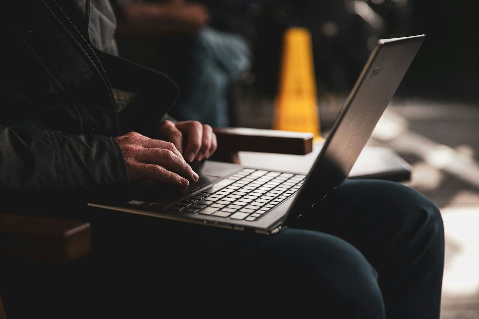 Close-up of a person’s hands typing on a silver laptop keyboard while seated, with a yellow cone and blurred street background behind them.